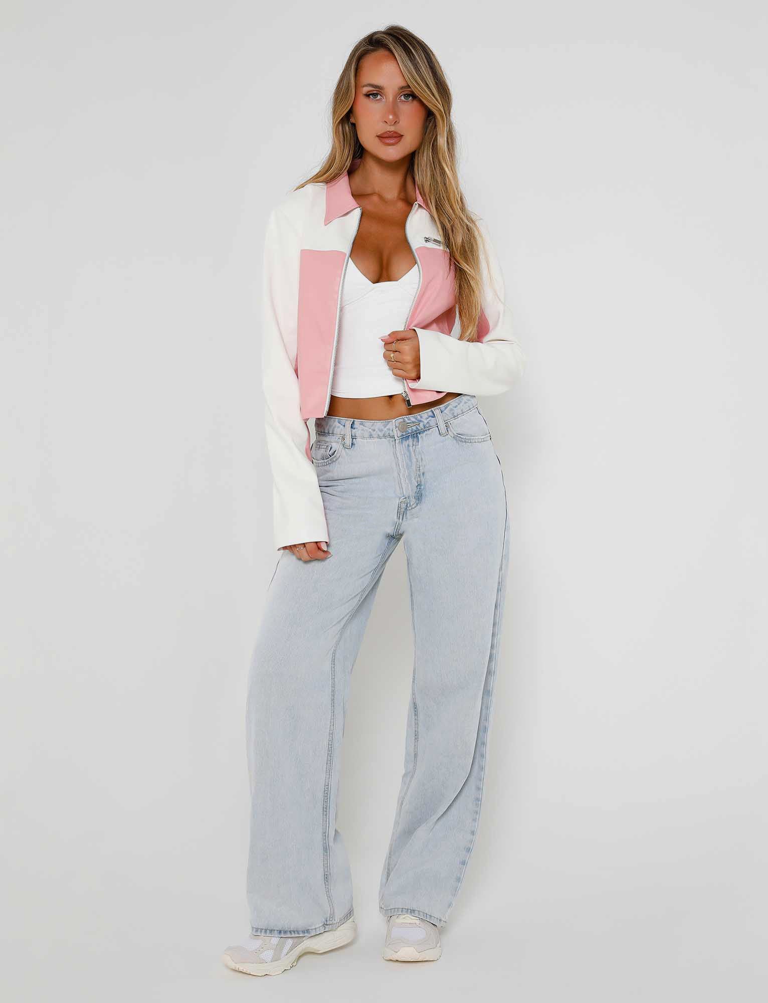 A woman with long, blonde hair stands confidently against a plain white background. She wears a white cropped tank top, light blue wide-leg jeans, white sneakers, and the TIGERMIST DELIA JACKET - PINK in pink and white, which she grasps with one hand, slightly exposing her midriff.