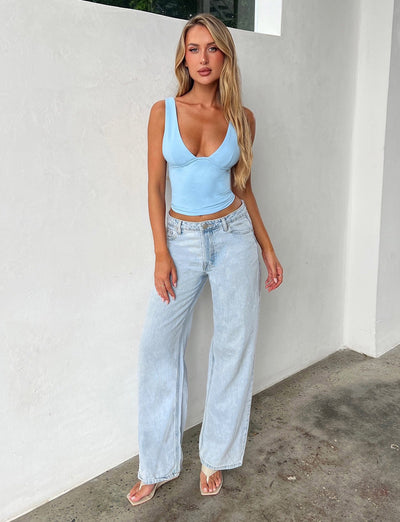 A woman stands against a white wall, showcasing the TIGERMIST LUNARIA TOP - BLUE : BABY BLUE, paired with light wash, wide-leg jeans. Her long blonde hair flows freely as she wears white sandals and poses with one hand on her hip and a relaxed expression.