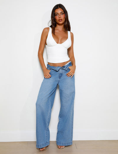 A woman stands against a white wall, wearing the LUNARIA TOP - WHITE from TIGERMIST and high-waisted, wide-leg blue jeans. She has her hands in her pockets and her dark hair is styled in loose waves. She is looking directly at the camera with a neutral expression.