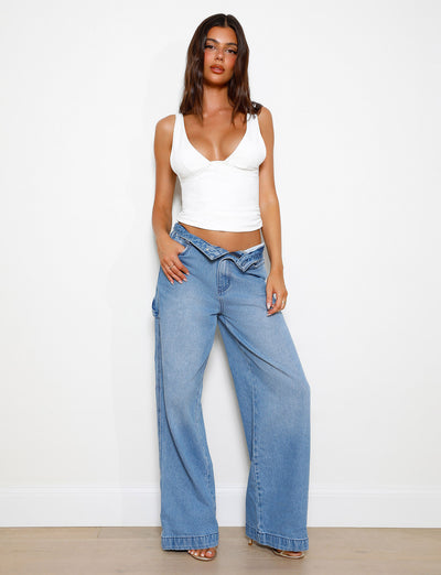 A woman is standing against a blank wall, wearing the LUNARIA TOP - WHITE by TIGERMIST and loose-fitting blue jeans. She has long, dark hair and is posing with one hand resting on her thigh and the other in her pocket. She is wearing neutral-colored high heels.
