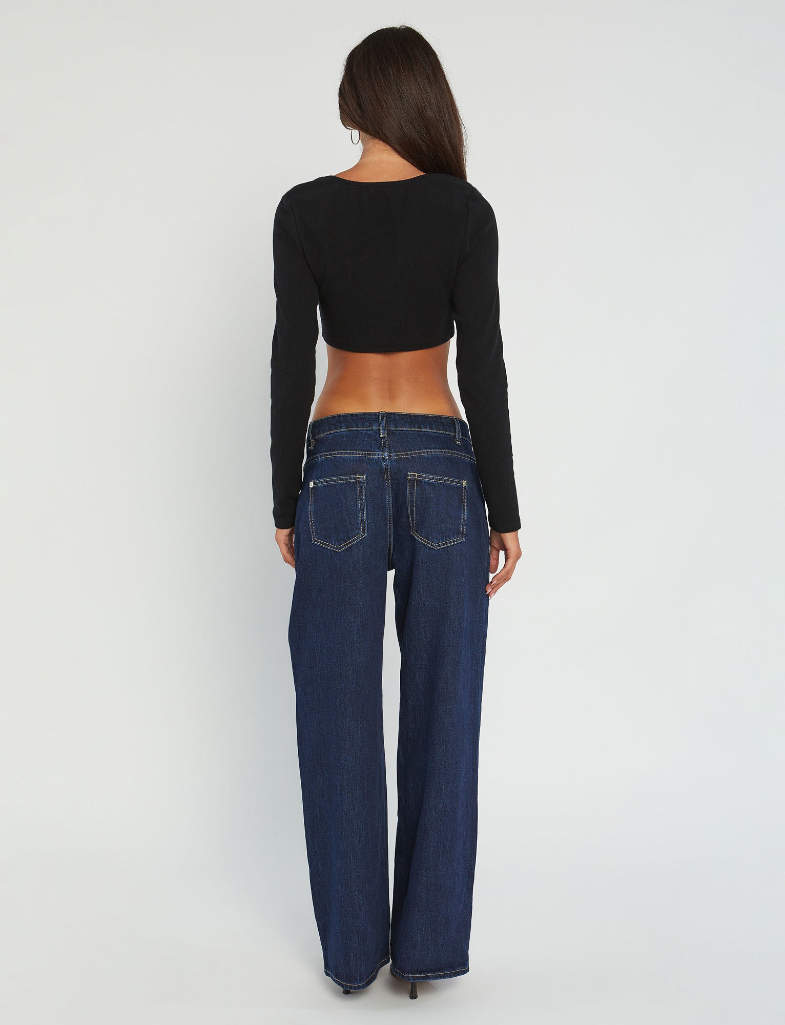 A person with long dark hair, seen from the back, is showcasing a chic MARYANNE TOP - BLACK by TIGERMIST and dark blue low-rise jeans with visible back pockets. The background is light gray.