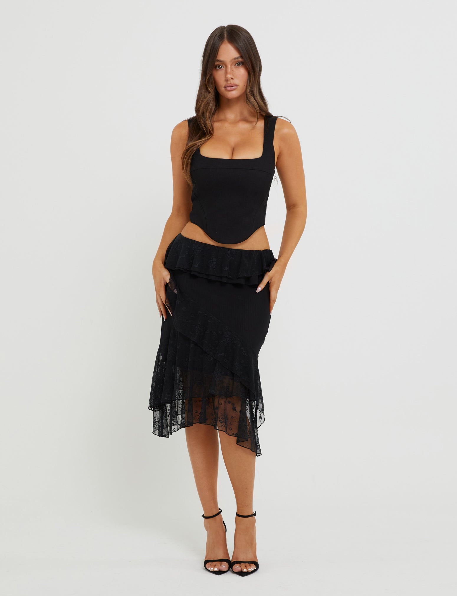A woman stands against a plain white background, wearing the SULLIVAN TOP - BLACK from TIGERMIST, which features a square neckline. She is also dressed in a fitted, asymmetrical black skirt with lace detailing by Tiger Mist. Her long, wavy brown hair cascades down her shoulders, and she is wearing black high-heeled sandals.