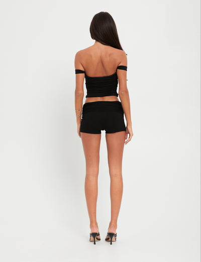 A person with long dark hair, facing away from the camera, is dressed head to toe in TIGERMIST. They are wearing the black SAFAA TOP along with matching black shorts and high heels against a plain white background.