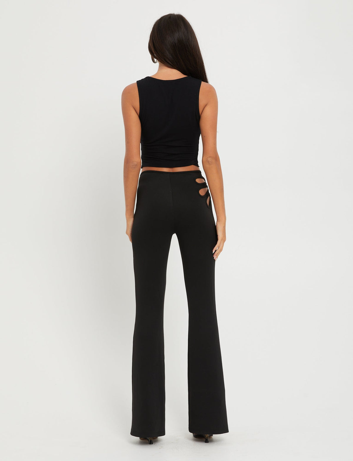 A woman with long dark hair stands with her back to the camera, wearing a TIGERMIST KATO TOP - BLACK and high-waisted black pants with a cut-out design on the side of the waist. She is standing against a plain white background.