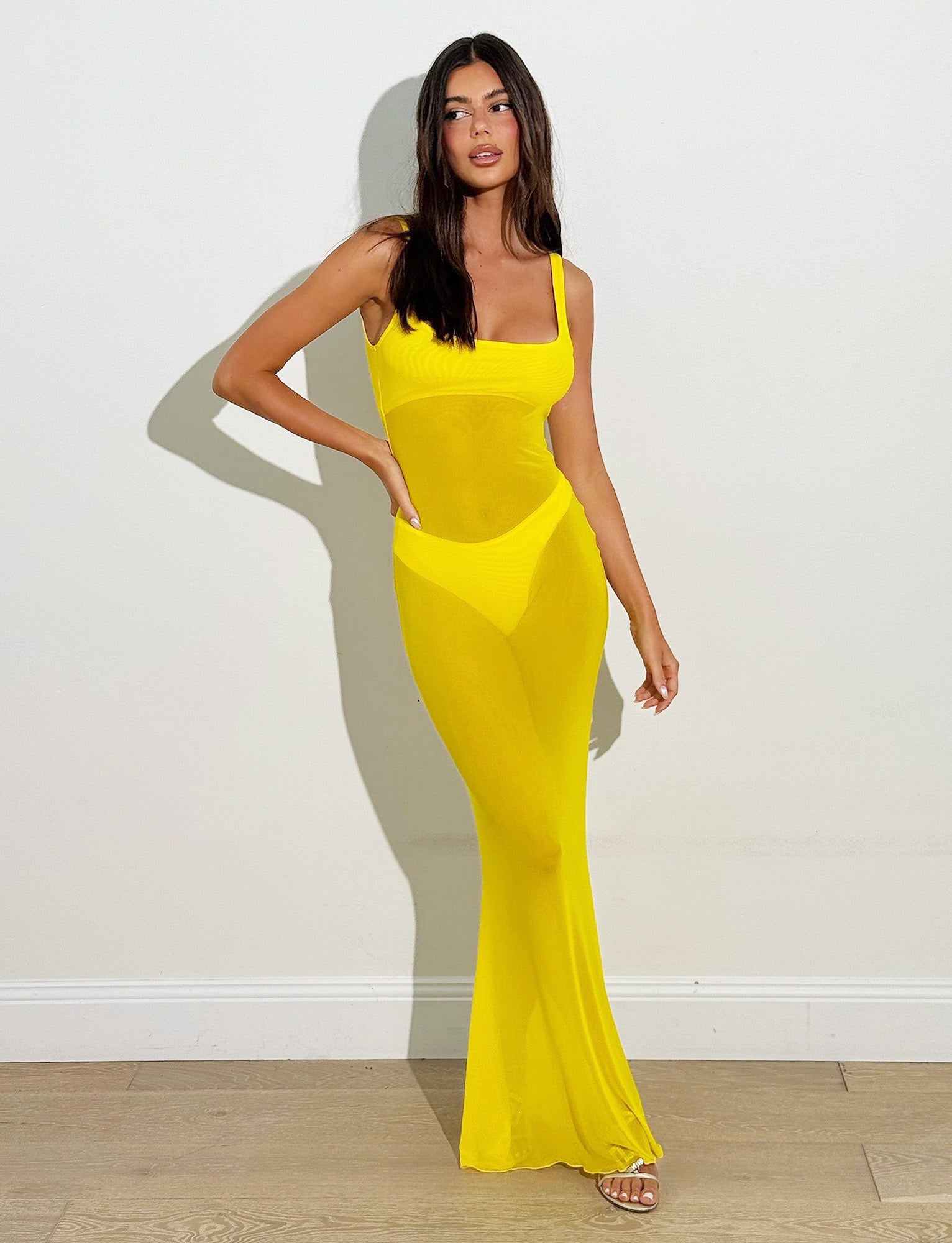 A woman with long dark hair stands against a plain white wall, wearing the fitted SAMIR DRESS by TIGERMIST. The bright yellow ensemble features cut-out details at the sides and midriff. With one hand on her hip and the other hanging by her side, she poses confidently on wooden flooring.