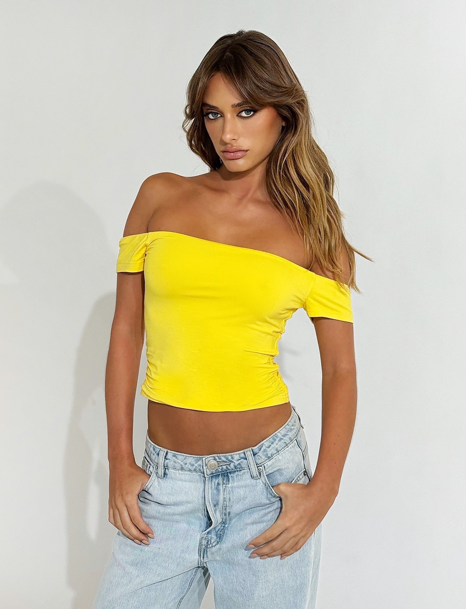 A person with long, wavy hair is wearing a bright yellow CAIDEN TOP from TIGERMIST and light blue jeans. They are standing against a plain white background, with one hand resting on their hip and the other hanging by their side.