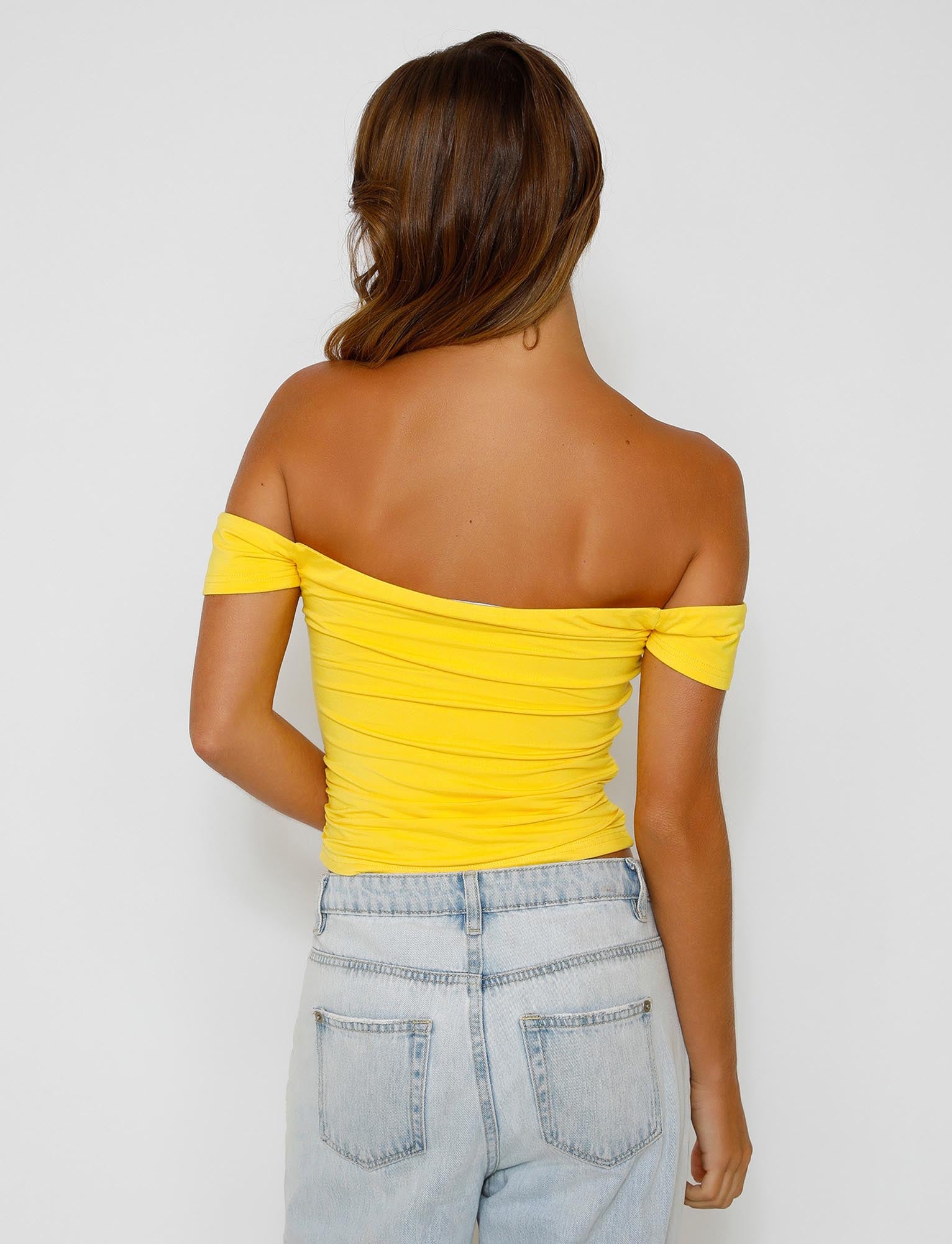 A person with long hair is standing with their back facing the camera, showcasing a stylish TIGERMIST CAIDEN TOP in bright yellow, paired with light blue jeans. The background is plain white.