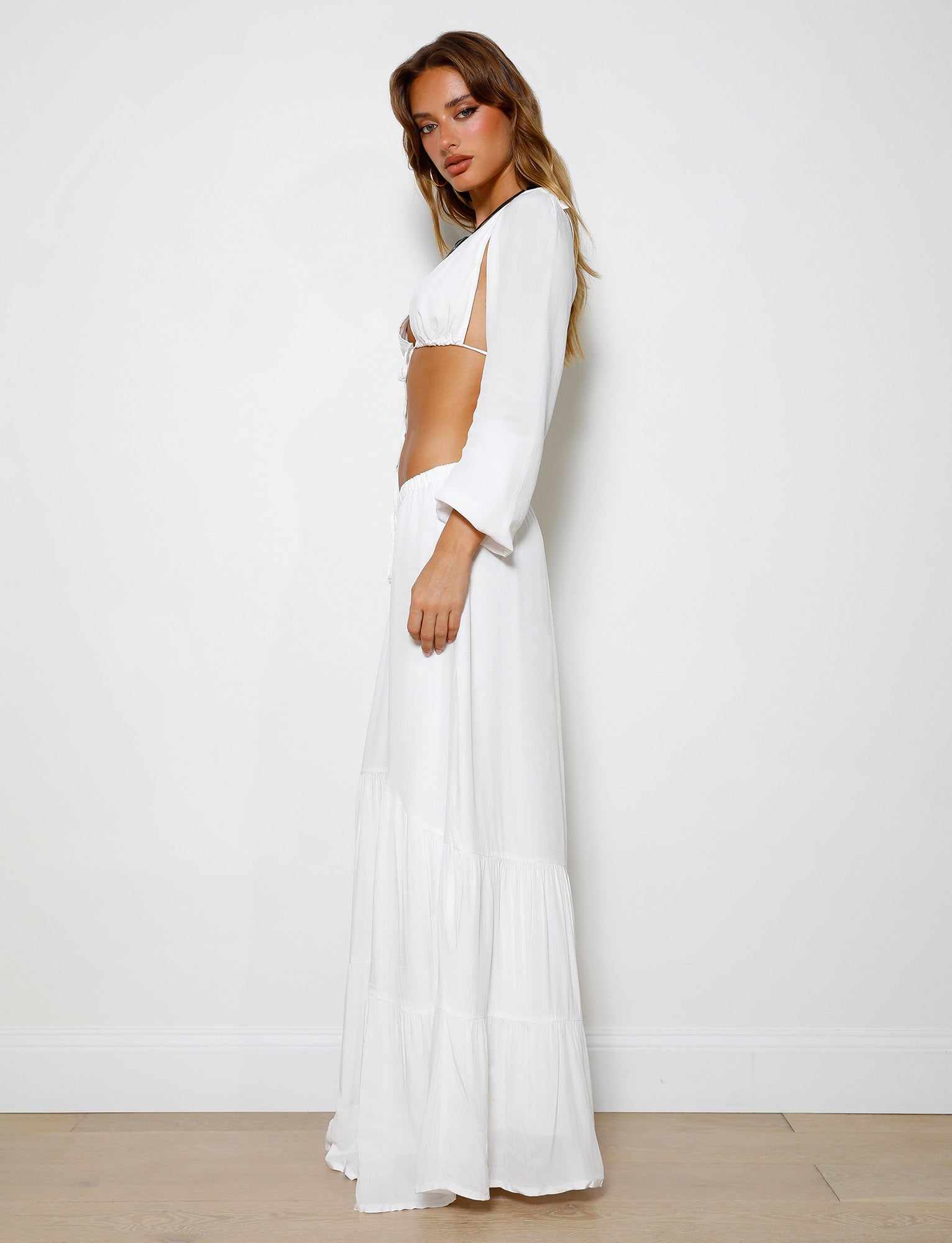 A woman stands against a plain white background, wearing the PHILLIPA SKIRT - WHITE by TIGERMIST with long sleeves and side cutouts at the waist. She is looking directly at the camera with a neutral expression, her blonde hair cascading over her shoulders.
