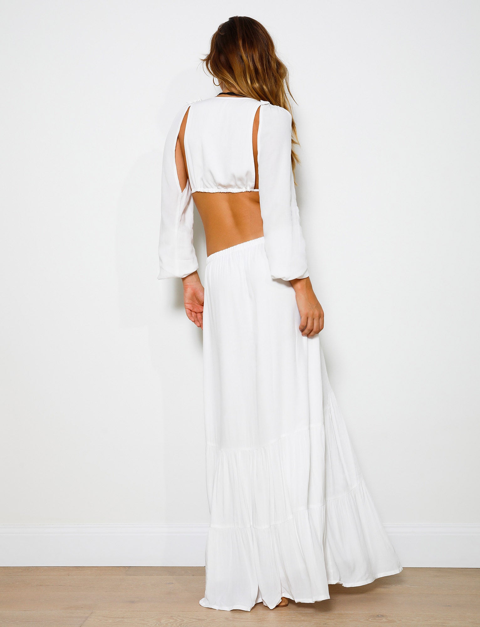 A woman stands against a white wall, facing away from the camera. She is wearing the PHILLIPA SKIRT - WHITE from TIGERMIST with cutout details on the back and sleeves. Her long, wavy hair falls over her shoulders and she stands on a light-colored wood floor.