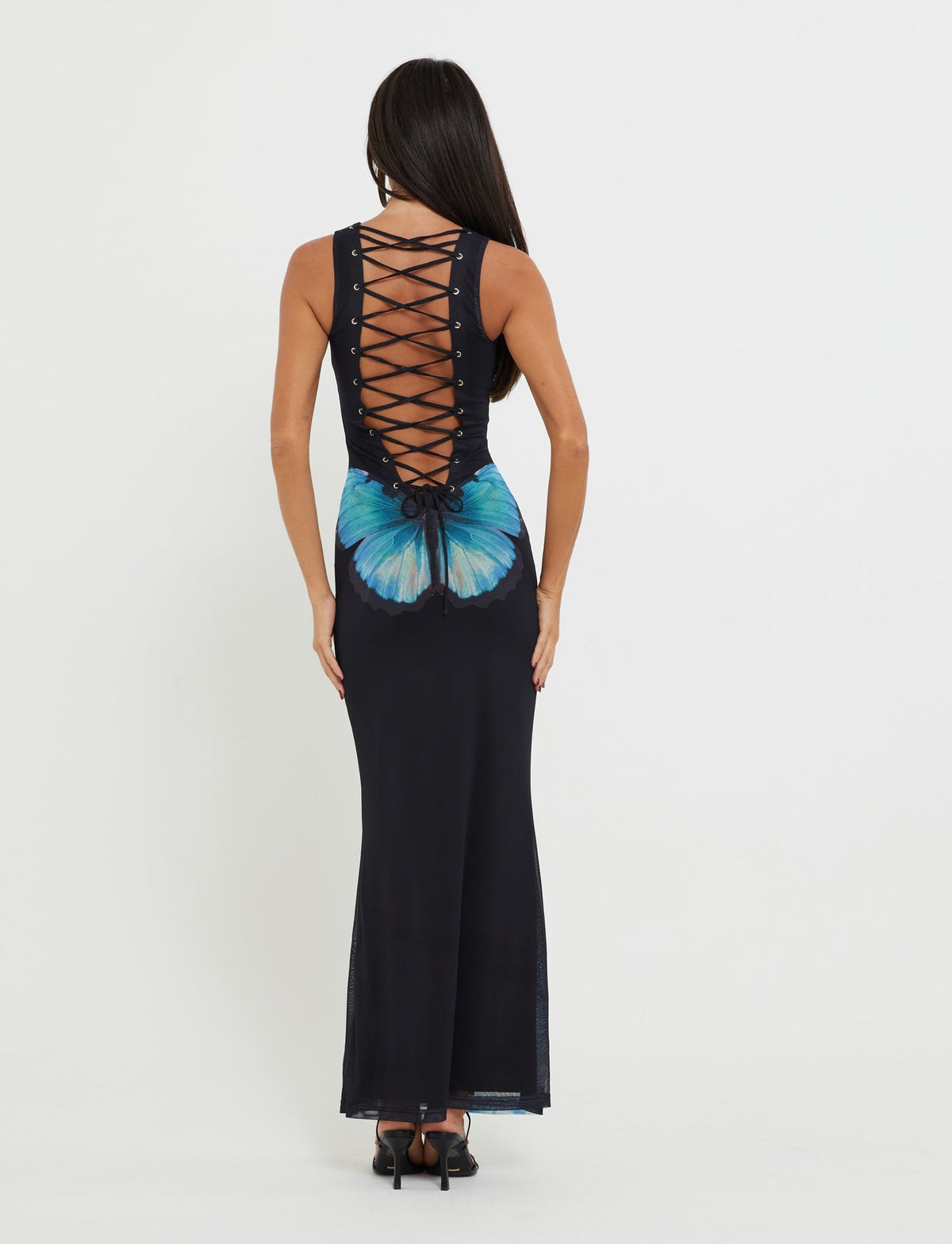 A woman wearing the ZAHRAA DRESS - BLACK : BUTTERFLY by TIGERMIST, a sleeveless, floor-length black dress with an open back, adorned with crossed lace-up details and a large butterfly design around the lower back and hips area. She stands with her back to the camera against a plain white background.