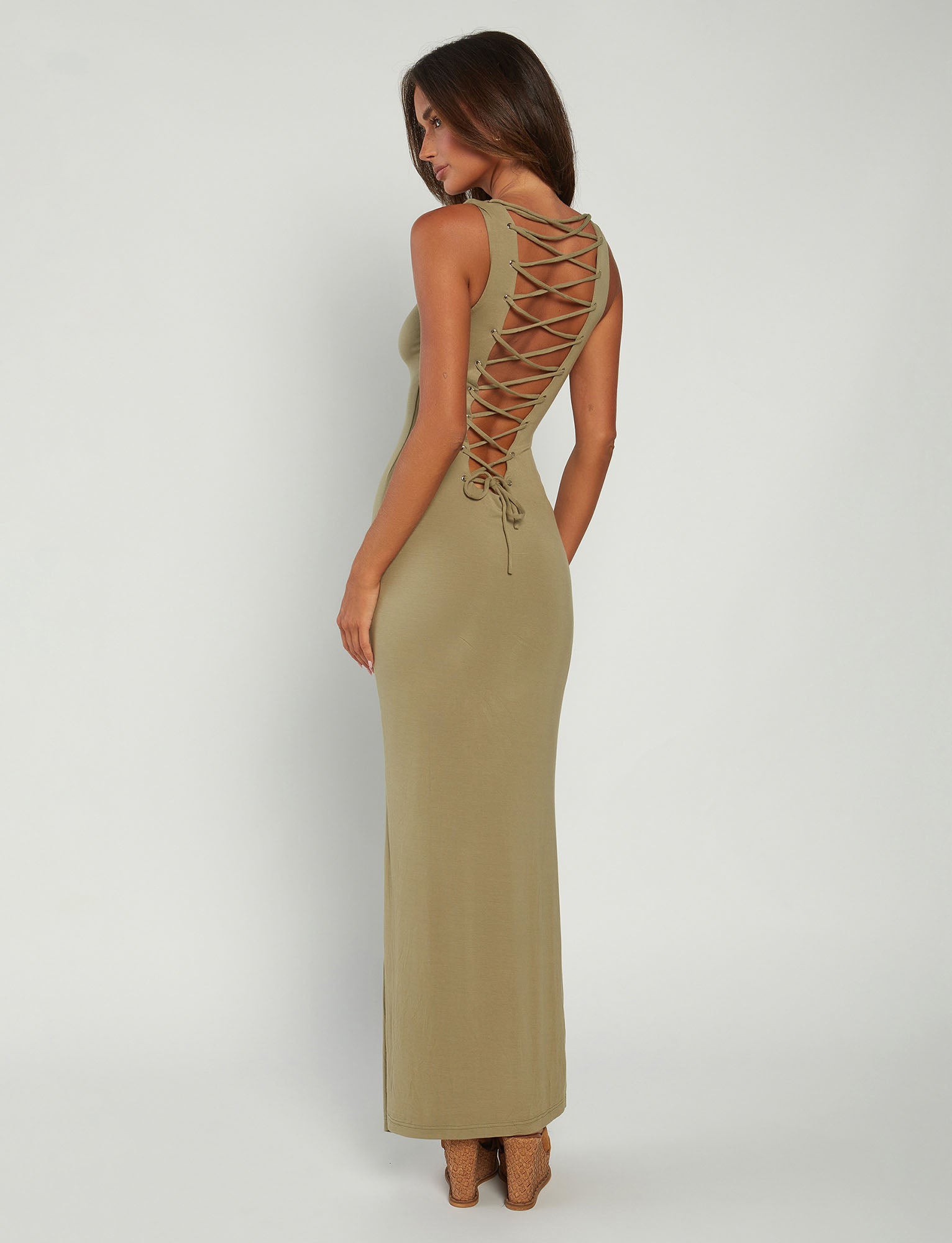A woman with long brown hair stands facing sideways and looks backward. Clad in a stylish TIGERMIST ZAHRAA Dress in moss green, featuring crisscross lacing down the back, she pairs it effortlessly with high-heeled, open-toed shoes. The background is plain white.