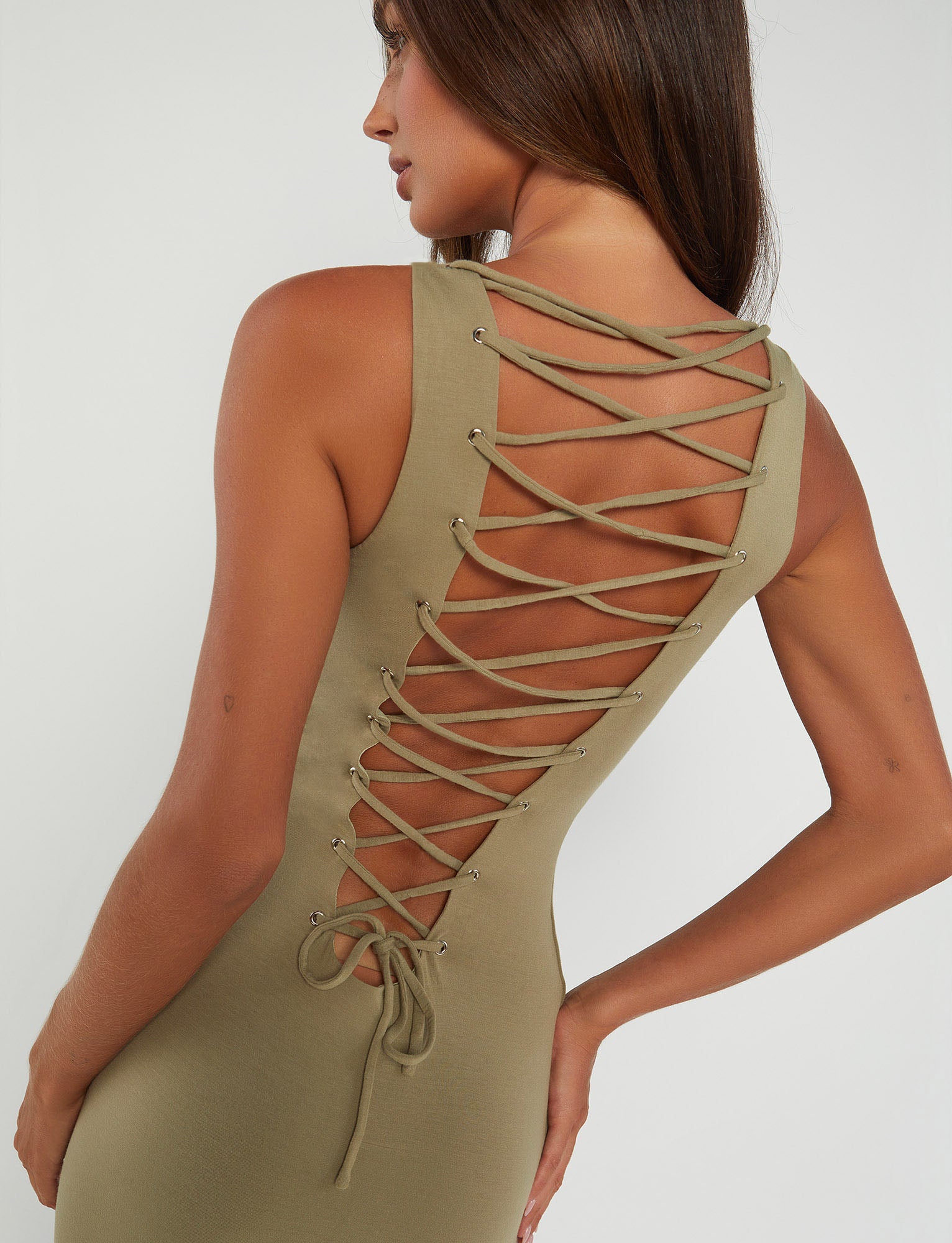 A woman is seen from the back, wearing a Moss Green Zahraa Dress by TIGERMIST, featuring a distinctive crisscross lace-up design running from the shoulders to the lower back. Her long brown hair falls over one shoulder as she stands against a plain white background.