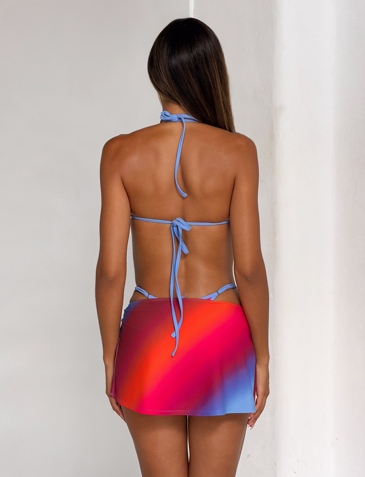 A woman with straight brown hair stands against a white wall, her back to the camera, wearing the TIGERMIST DIANA BIKINI TOP in Cielo Ombre, a vibrant gradient halter-neck style inspired by an ombre stripe pattern.