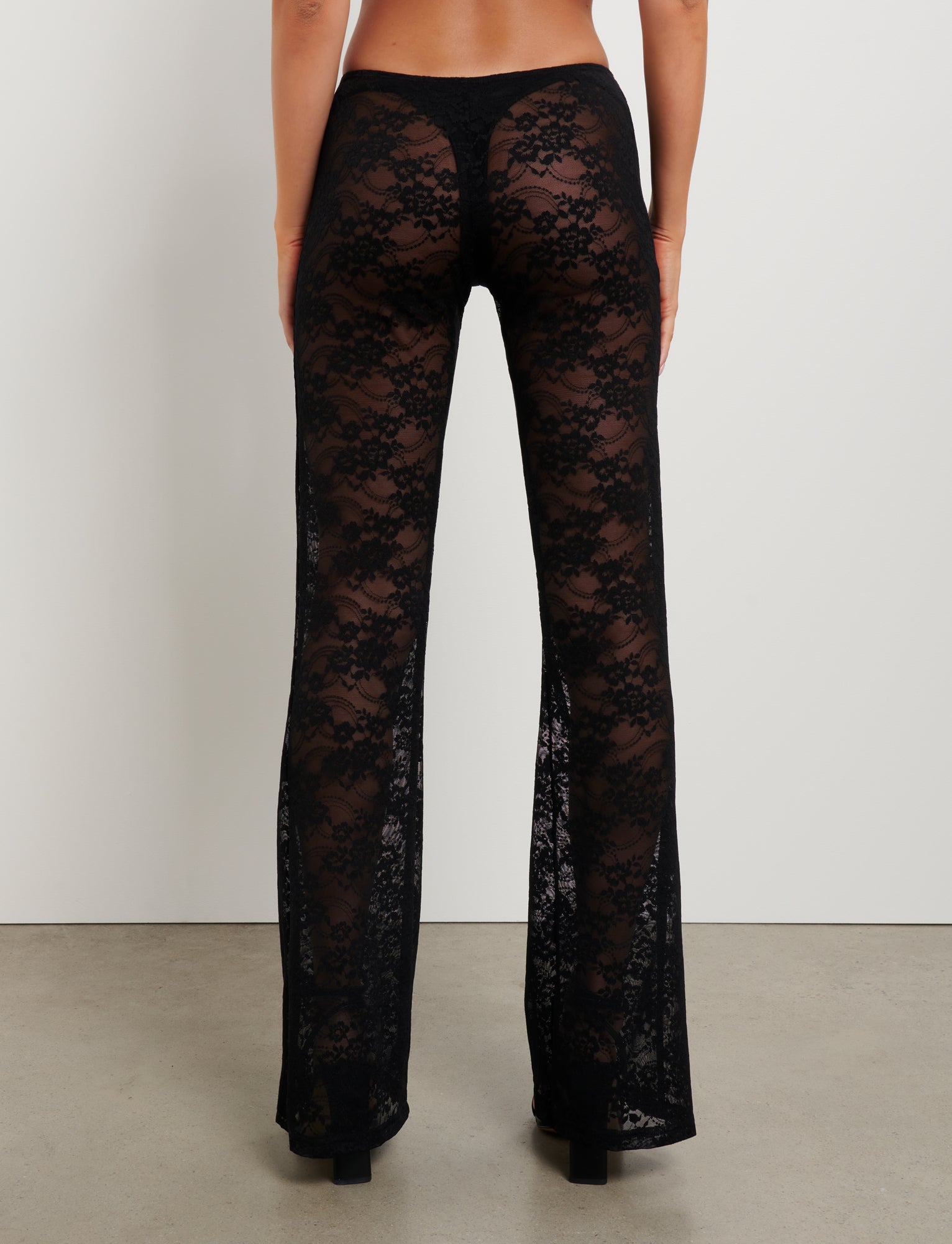 A person stands with their back to the camera, wearing the TIGERMIST MELANIE PANT in black lace, which reveals undergarments beneath. The pants feature a floral pattern and flare out slightly at the bottom. The person is standing against a plain white wall on a concrete floor.