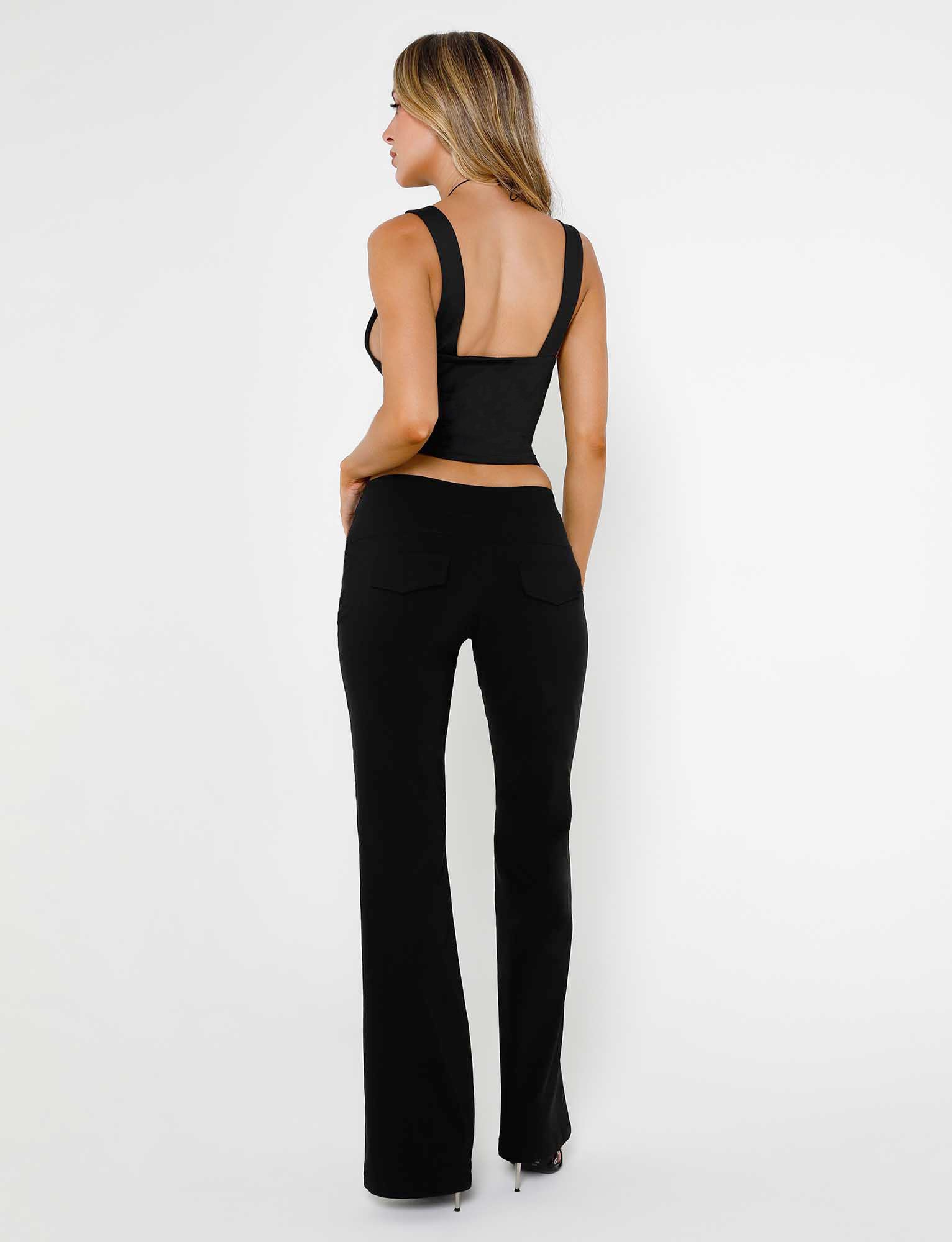 A woman with long hair, standing and facing away from the camera, is wearing the LUNARIA TOP - BLACK from TIGERMIST. She pairs it with black flared pants and heels, all set against a plain white background.