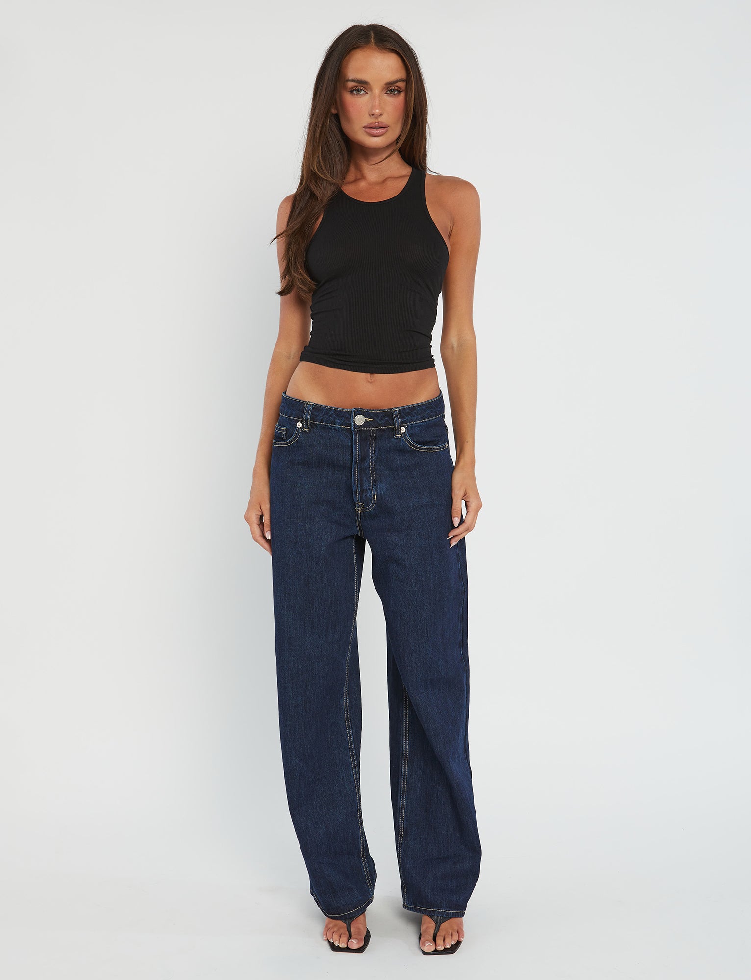 A woman stands against a plain white background, wearing the Tiger Mist SOMMER TANK in black and loose-fitting dark blue jeans. She has long, straight hair and is wearing black sandals. Her arms are relaxed by her sides.