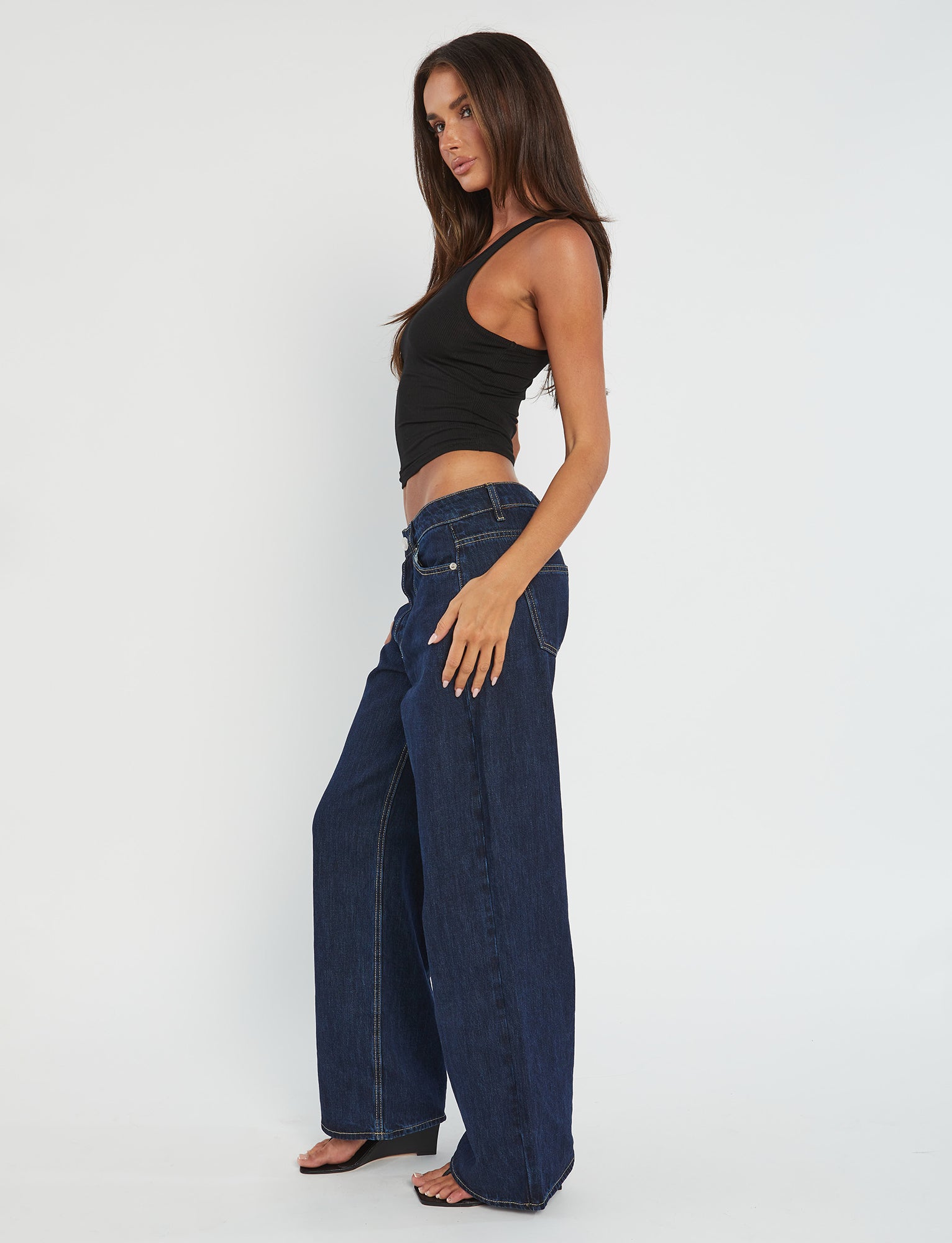 A person with long, dark hair is wearing the SOMMER TANK - BLACK from TIGERMIST and wide-leg dark blue jeans. They are standing sideways, facing left, with one hand by their side and the other slightly behind them, against a plain white background.