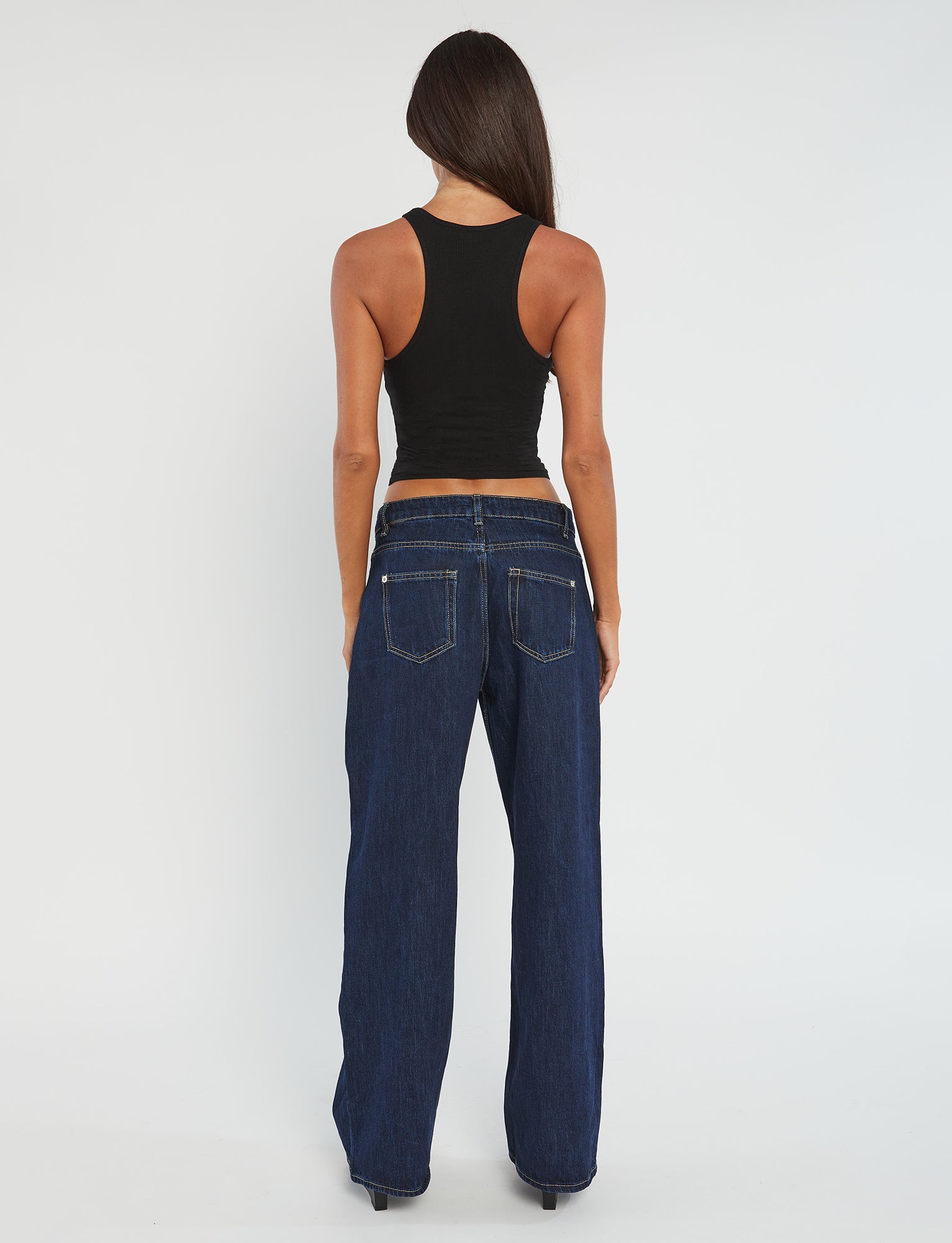 A woman with long dark hair stands with her back to the camera, dressed in a sleeveless SOMMER TANK - BLACK and dark blue wide-leg jeans from TIGERMIST. The background is plain white.