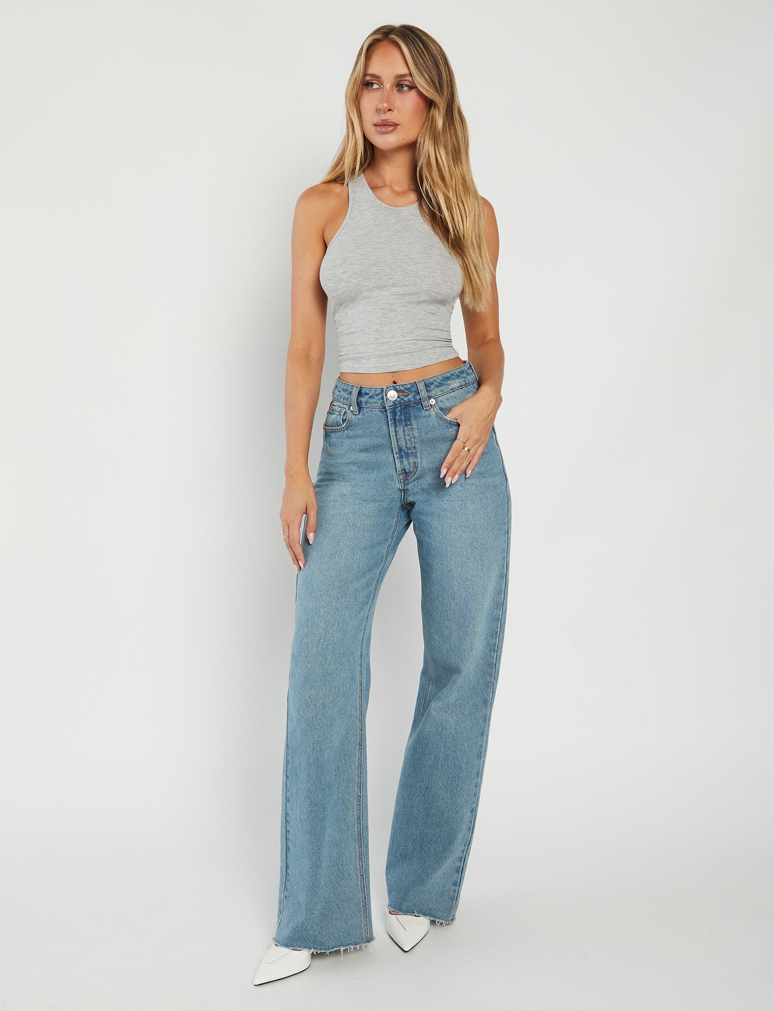 A woman with long, wavy blonde hair stands against a plain white background. Dressed in the SOMMER TANK - GREY and high-waisted blue jeans from TIGERMIST, she completes her look with white pointed-toe shoes. She has a relaxed expression, her left arm resting by her side while her right hand touches her hip.