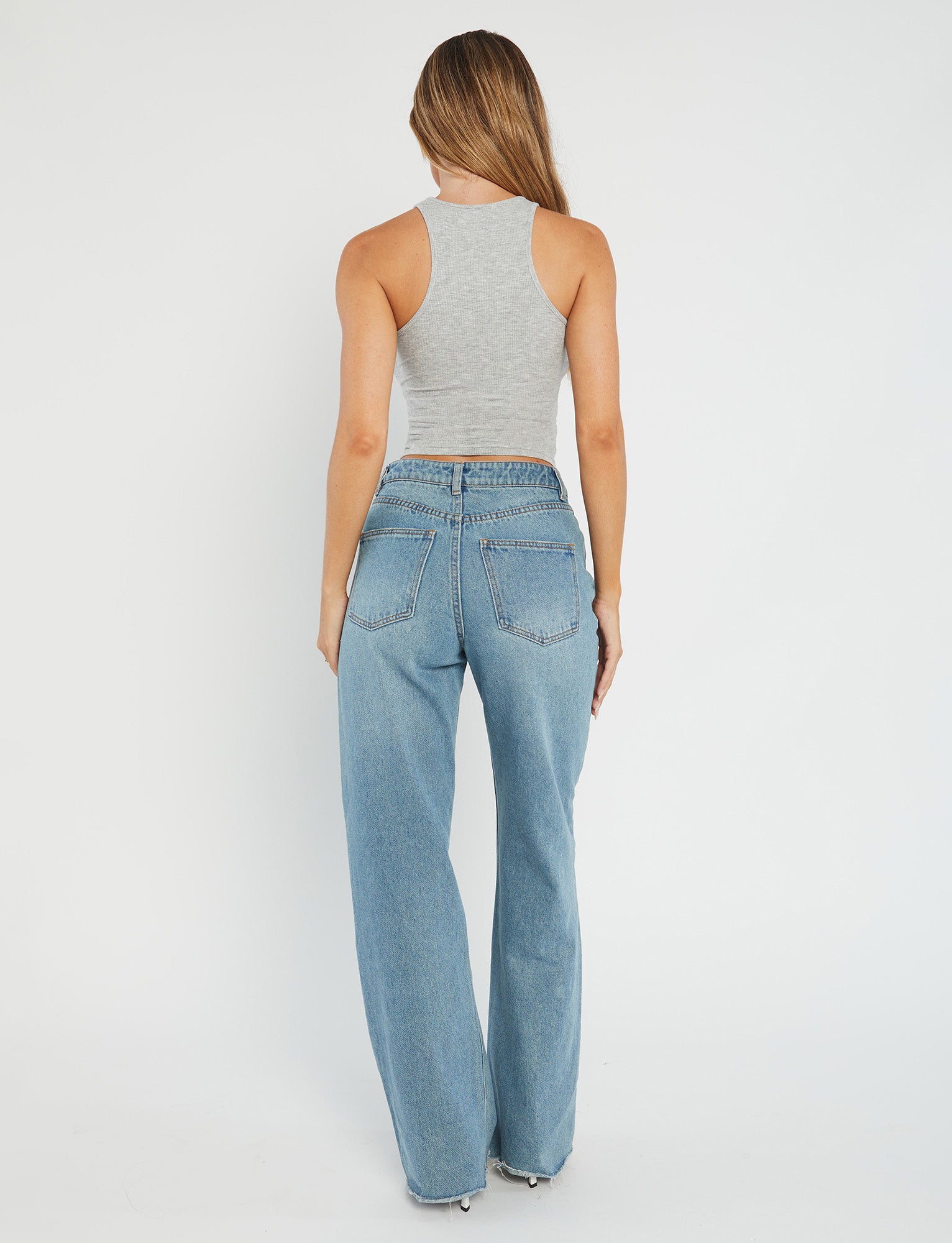 A person stands facing away from the camera, donning the SOMMER TANK in grey and high-waisted blue jeans by TIGERMIST. The background is a plain, light-colored wall. The individual has long, straight hair and exudes a relaxed posture.
