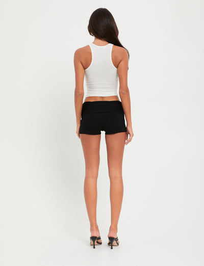 A person with long dark hair is standing with their back to the camera. They are wearing the TIGERMIST SOMMER TANK in white, black shorts, and black high-heeled shoes. The background is plain and white.