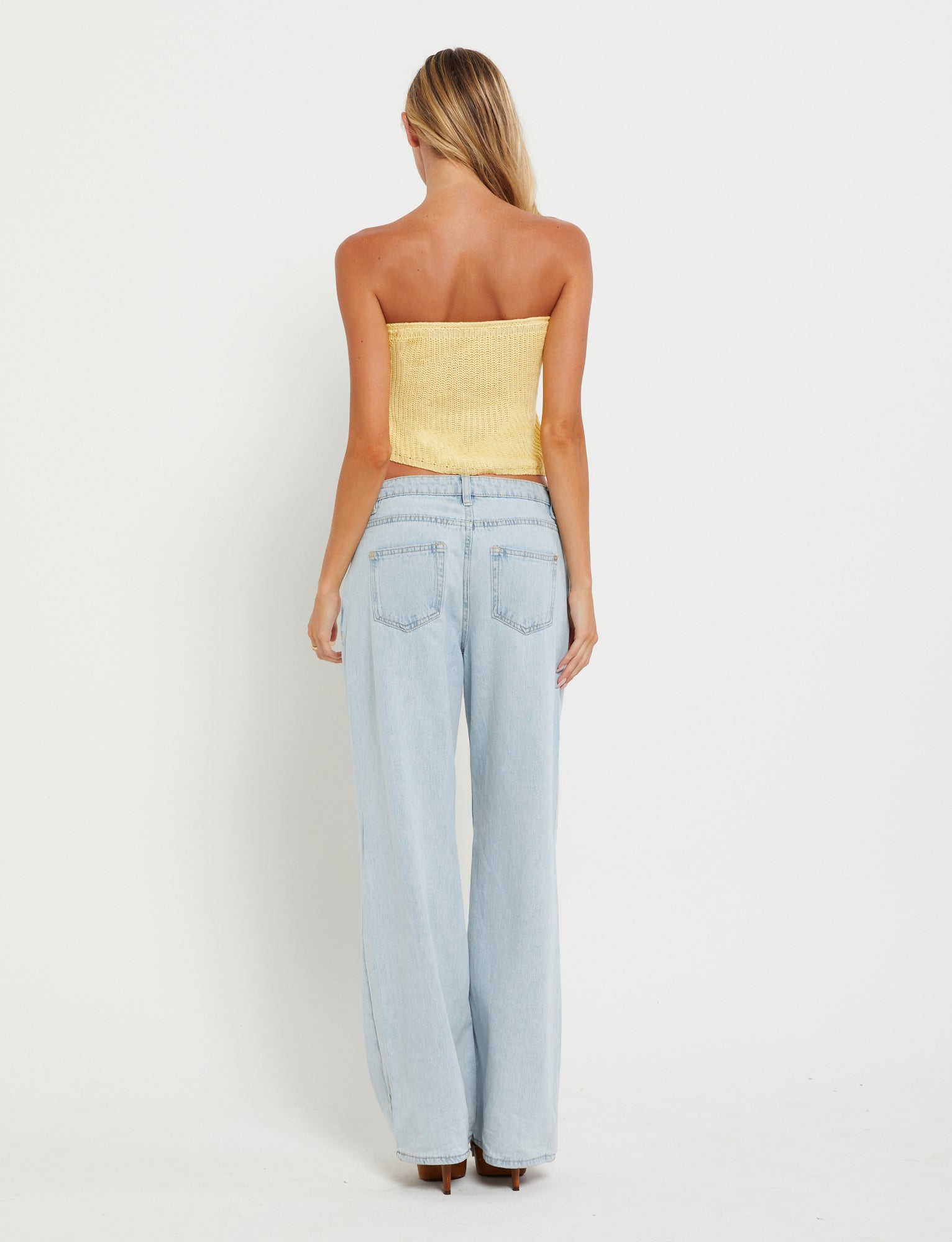 A woman with long blonde hair stands with her back to the camera, wearing a TIGERMIST MORAI TOP in Lemon and light blue high-waisted jeans against a plain white background.