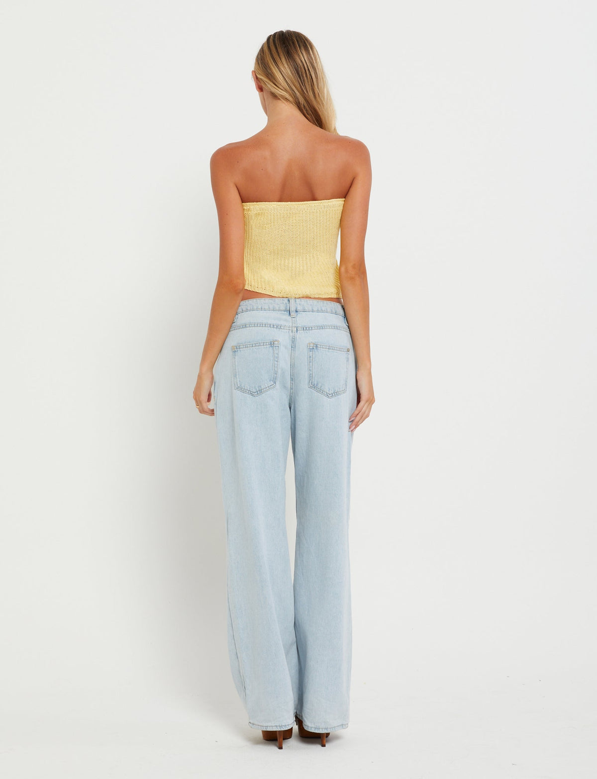 A woman with long blonde hair stands with her back to the camera, wearing a TIGERMIST MORAI TOP in Lemon and light blue high-waisted jeans against a plain white background.