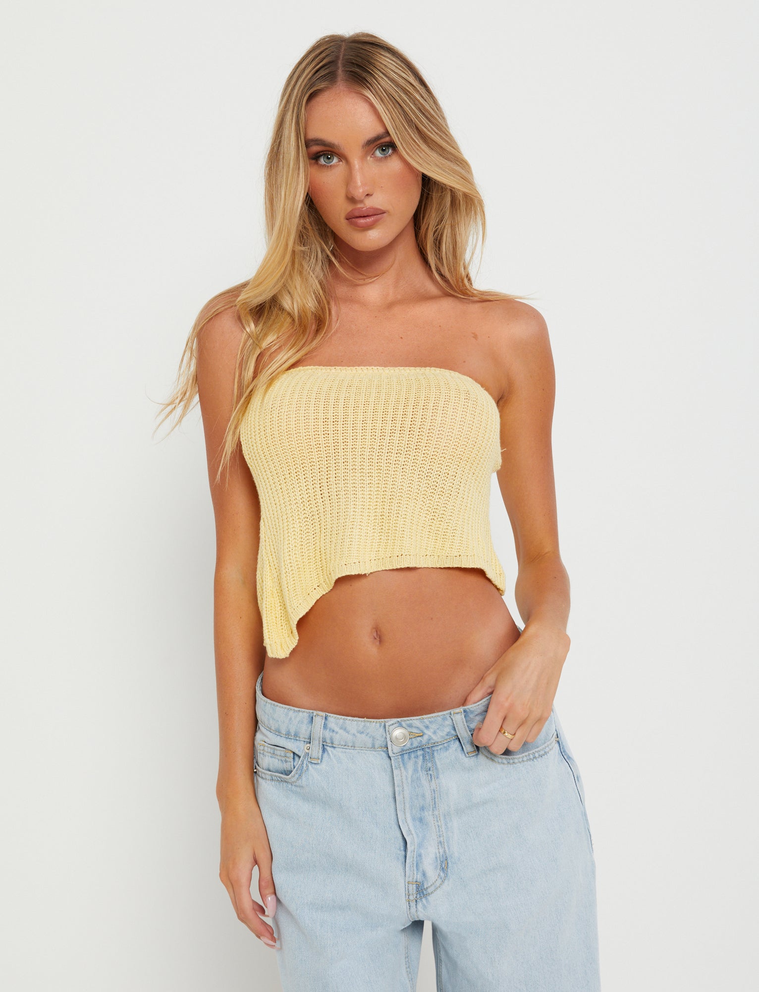 A blonde woman with long hair is wearing the MORAI TOP - YELLOW : LEMON, a strapless knit crop top from TIGERMIST, along with light blue, slightly oversized jeans. She stands in front of a plain white background, with her right hand resting on her hip.