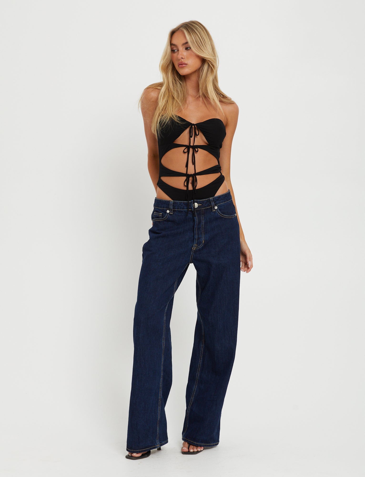 A blonde woman stands against a plain white background, wearing the KINERA BODYSUIT - BLACK from TIGERMIST and blue wide-leg jeans. She has her left hand resting on her thigh and her right hand lightly touching her upper arm.