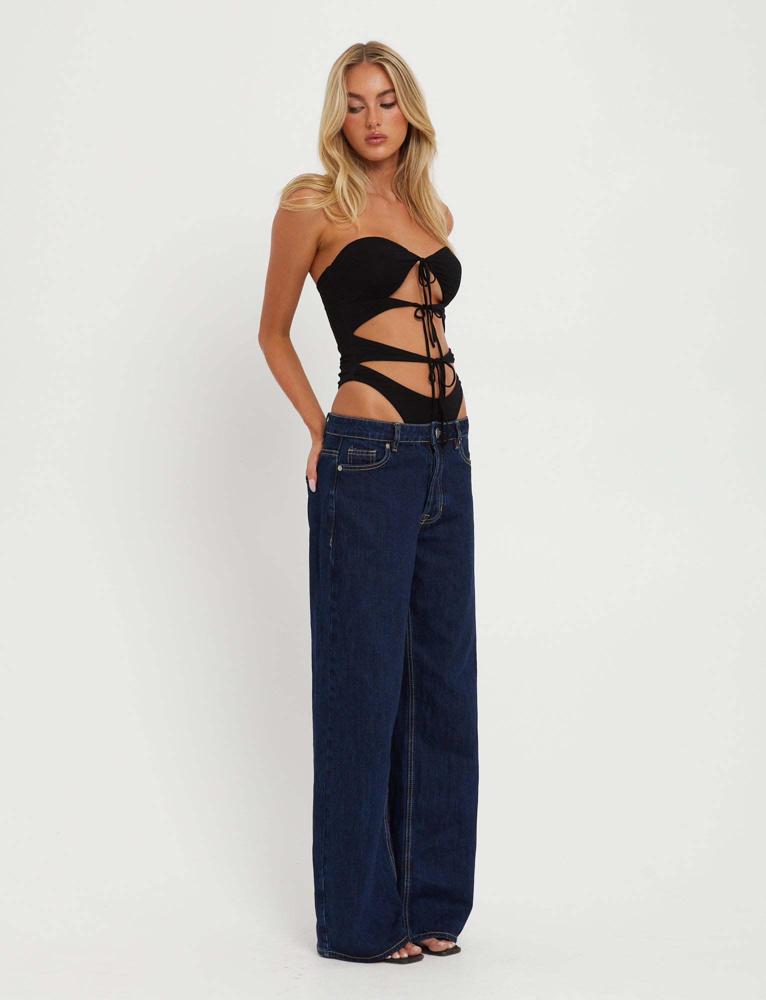 A woman with long blonde hair is modeling the KINERA BODYSUIT in black from TIGERMIST and high-waisted, wide-leg dark blue jeans. She stands against a plain white background, looking slightly down to her left with her hands in the front pockets of her jeans.