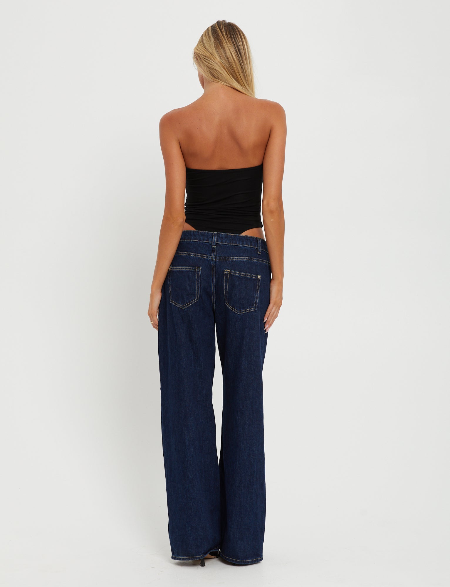 A person with long blonde hair, viewed from the back, is wearing the TIGERMIST KINERA BODYSUIT in black and high-waisted, wide-leg blue jeans. The background is plain white.