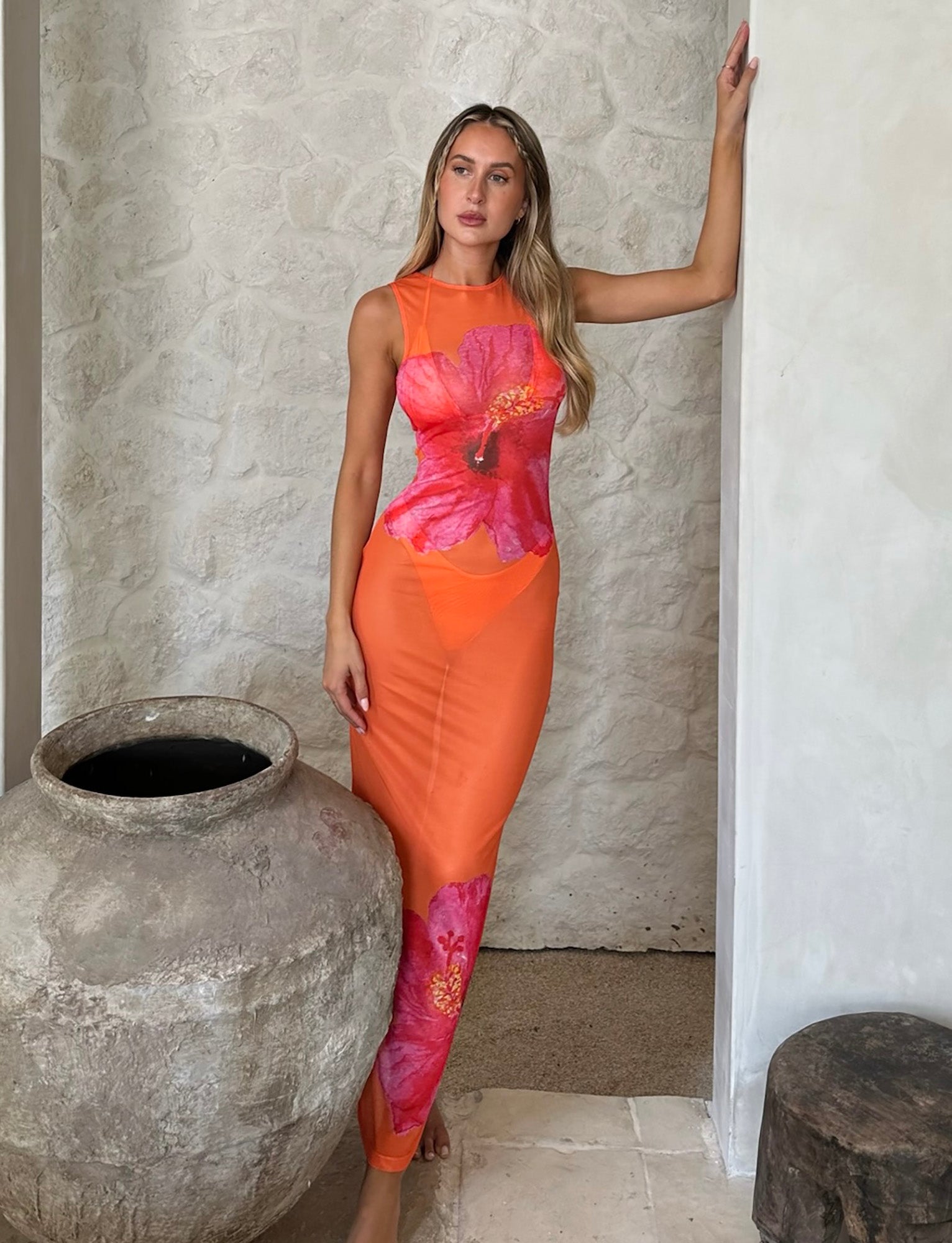 A woman with long blonde hair, wearing the JOZIE THREE PIECE DRESS in Orange Hibiscus from TIGERMIST, adorned with large pink flower prints, stands against a light-colored stone wall. She strikes a pose with one hand on her head and the other resting on a large clay pot beside her.