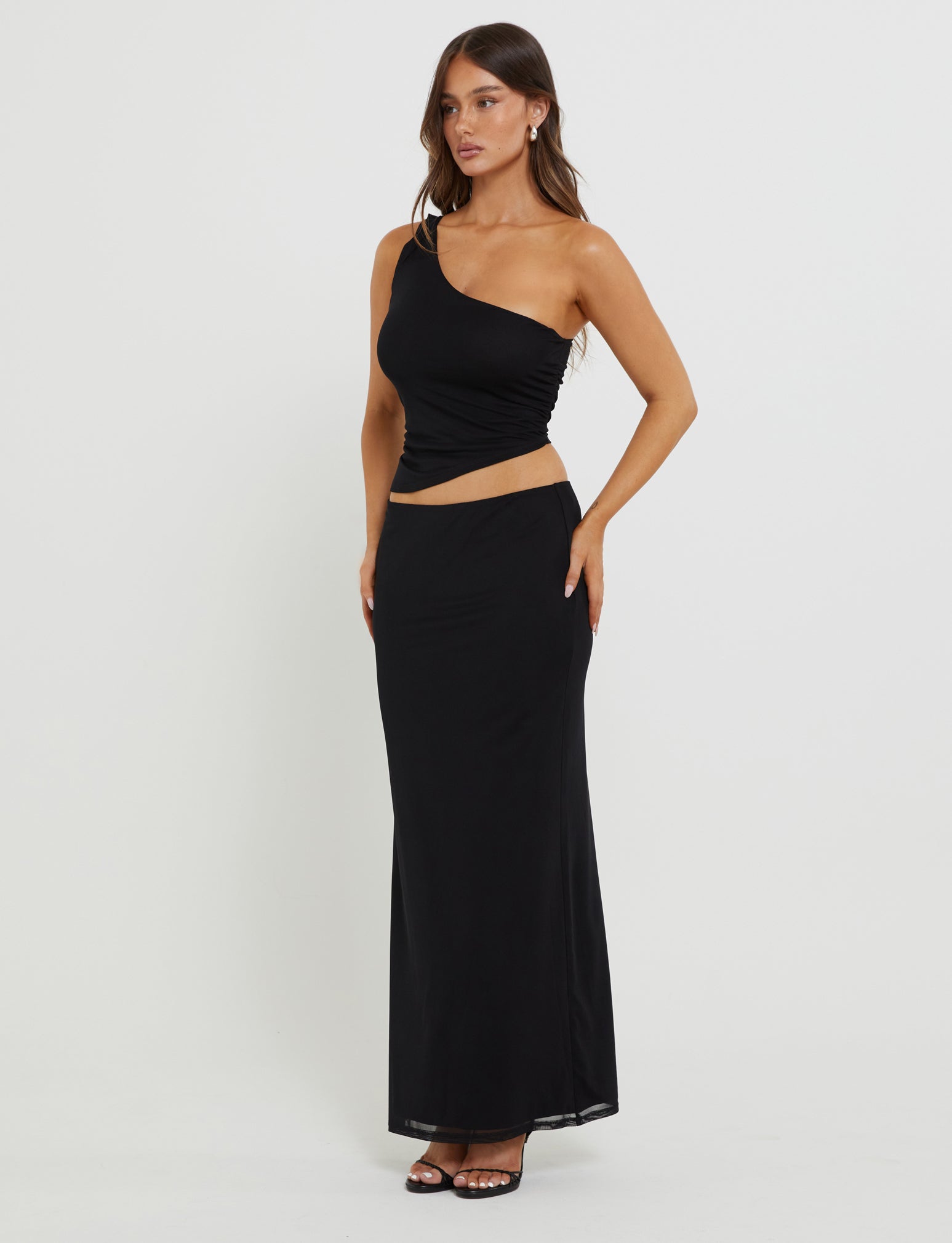 A woman stands against a plain white background, showcasing the DEANNA TOP - BLACK from TIGERMIST. She pairs the sleek black one-shoulder top with black open-toed heels and silver hoop earrings. Her long brown hair is styled down, and she gazes slightly to her left.