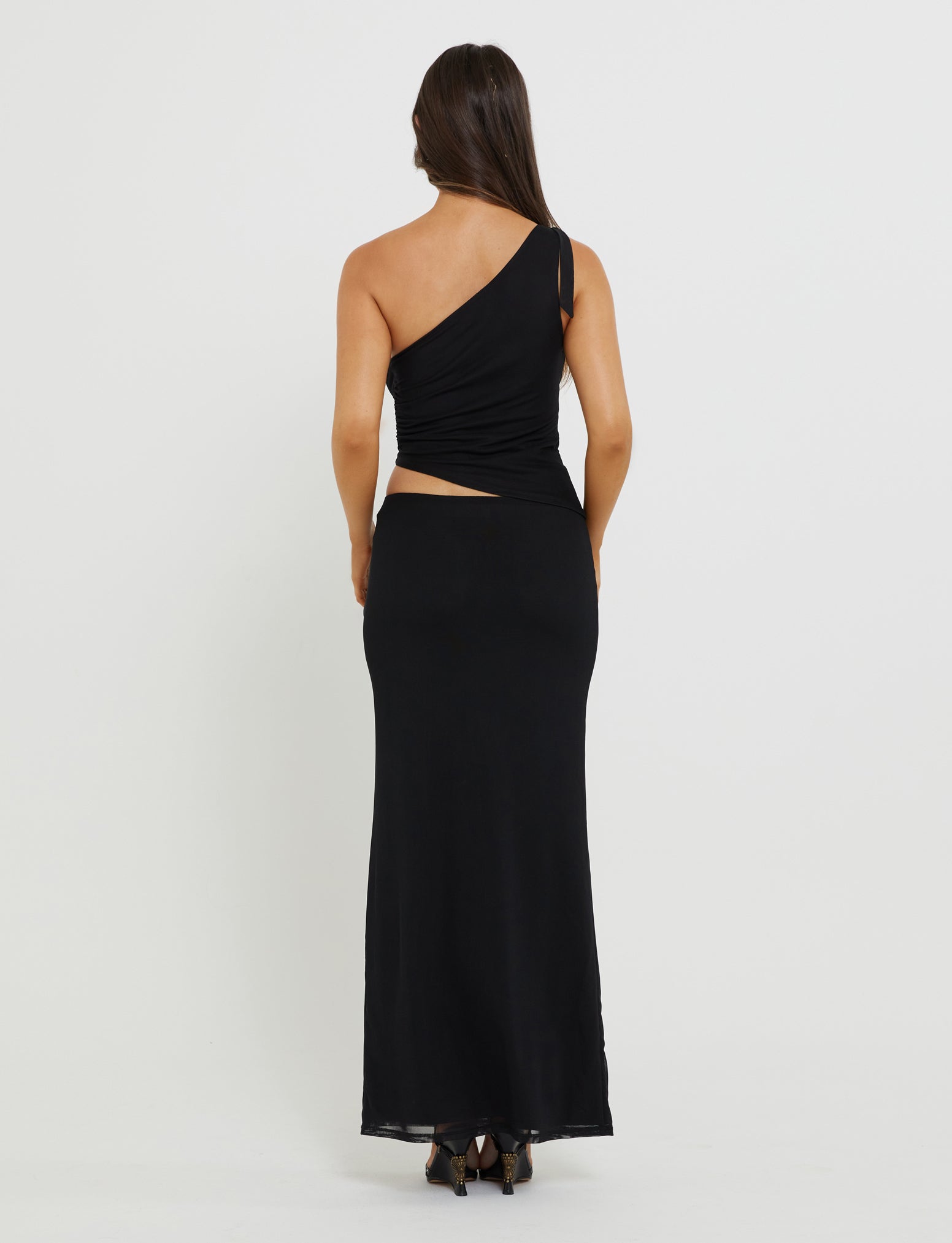 A person with long dark hair is standing with their back to the camera, showcasing a stunning Tiger Mist DEANNA TOP - BLACK paired with form-fitting pants and strappy black heels. The background is plain and white.