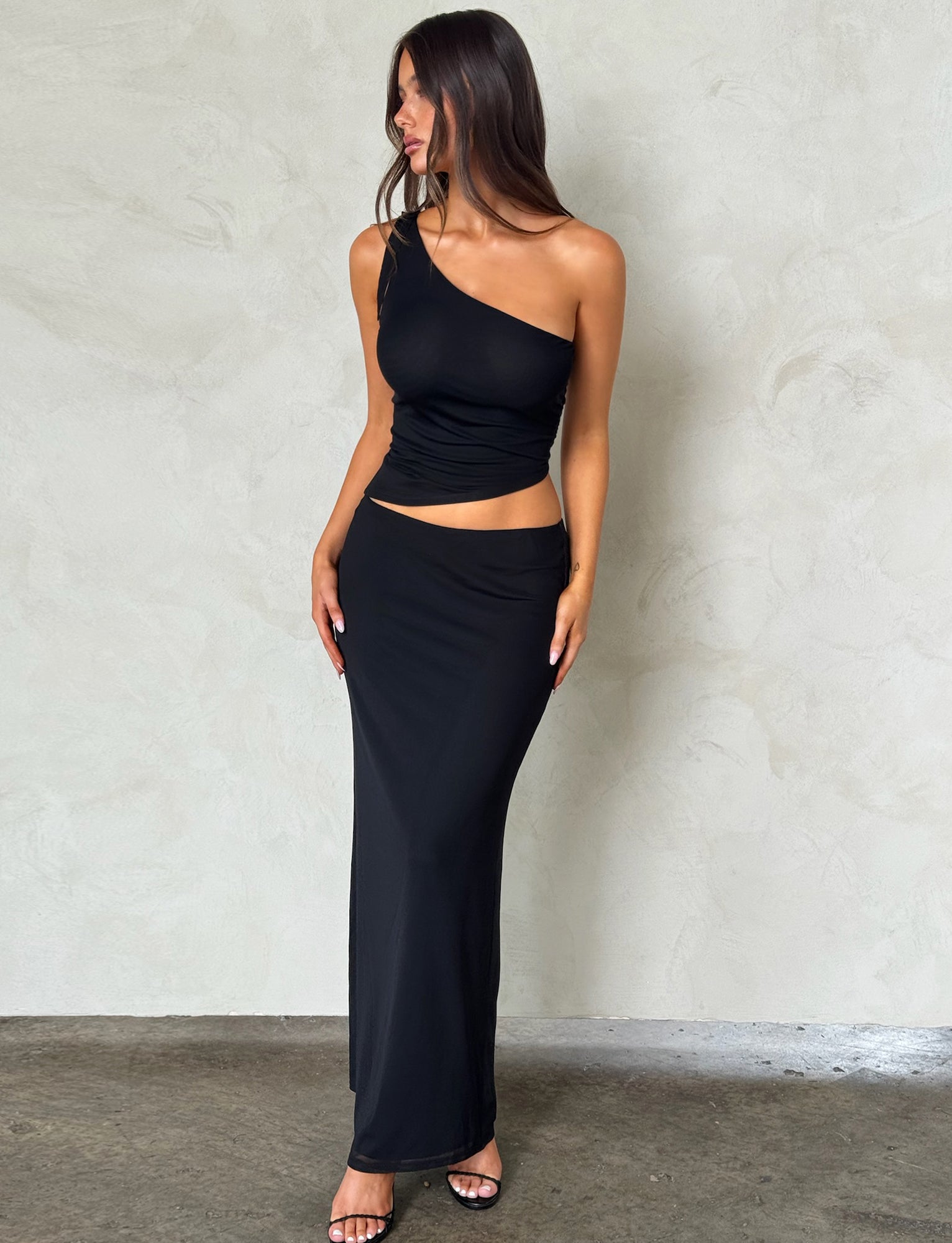 A woman with long, dark hair stands against a neutral background. She is wearing a one-shoulder, sleeveless black top from Tiger Mist, paired with the DEANNA MAXI SKIRT - BLACK from TIGERMIST and black open-toe heels. She has a relaxed and confident pose.