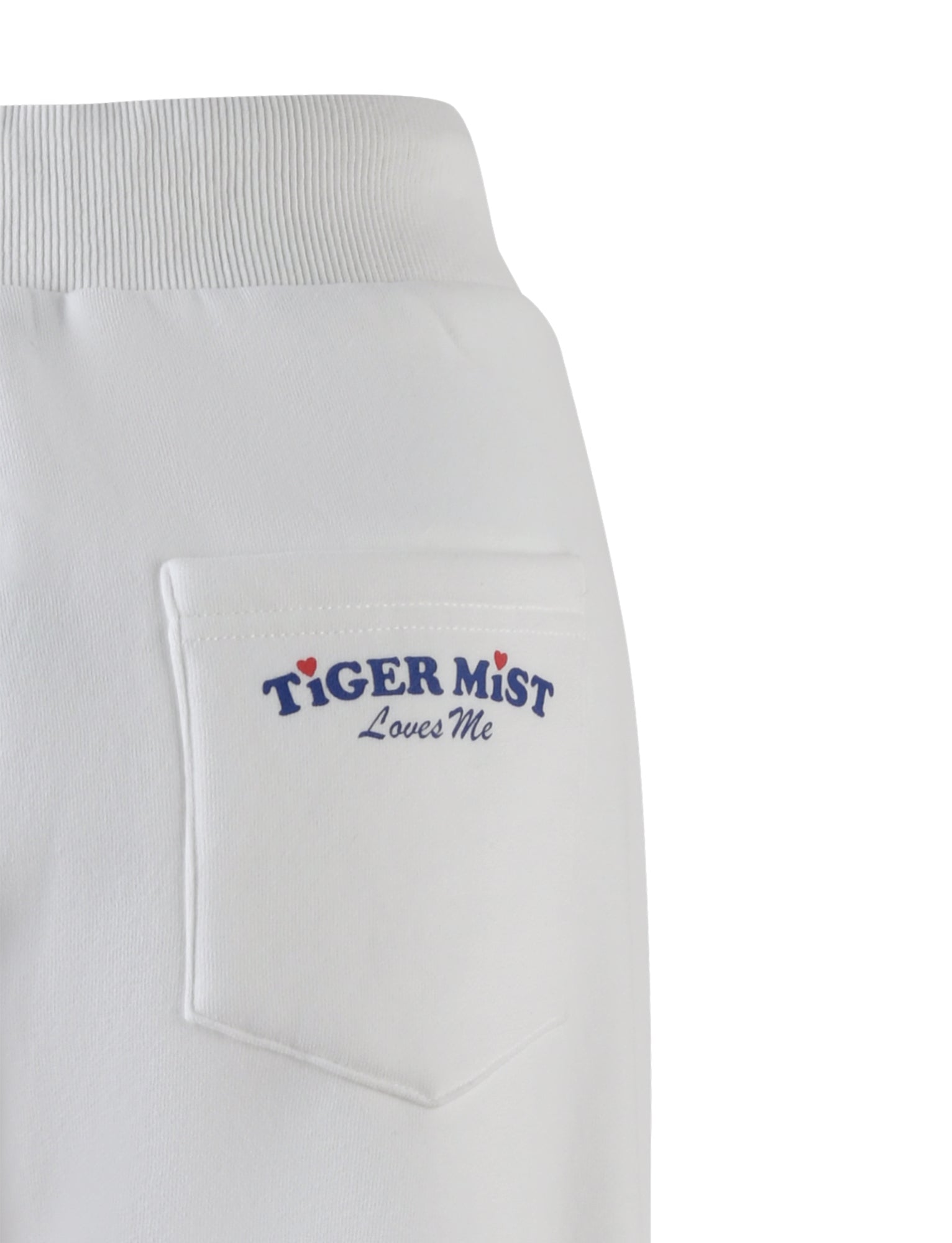Close-up image of the back pocket of the LUMEN PANT - WHITE by TIGERMIST, featuring the words "TIGER MIST Loves Me" printed in blue cursive lettering above the pocket. The ribbed waistband adds a comfortable touch, and two small red hearts dot the "I" in "TIGER" and "LOVES". An oversized trackpant silhouette completes the look.