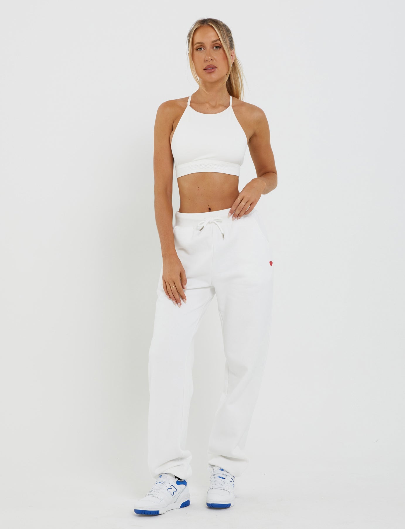 A woman stands against a white background wearing a white sports bra, the TIGERMIST LUMEN PANT in white, featuring a mid-rise waist with drawstring and an oversized trackpant silhouette, and white sneakers with blue accents. She has her left hand in her pocket and her right arm relaxed by her side.