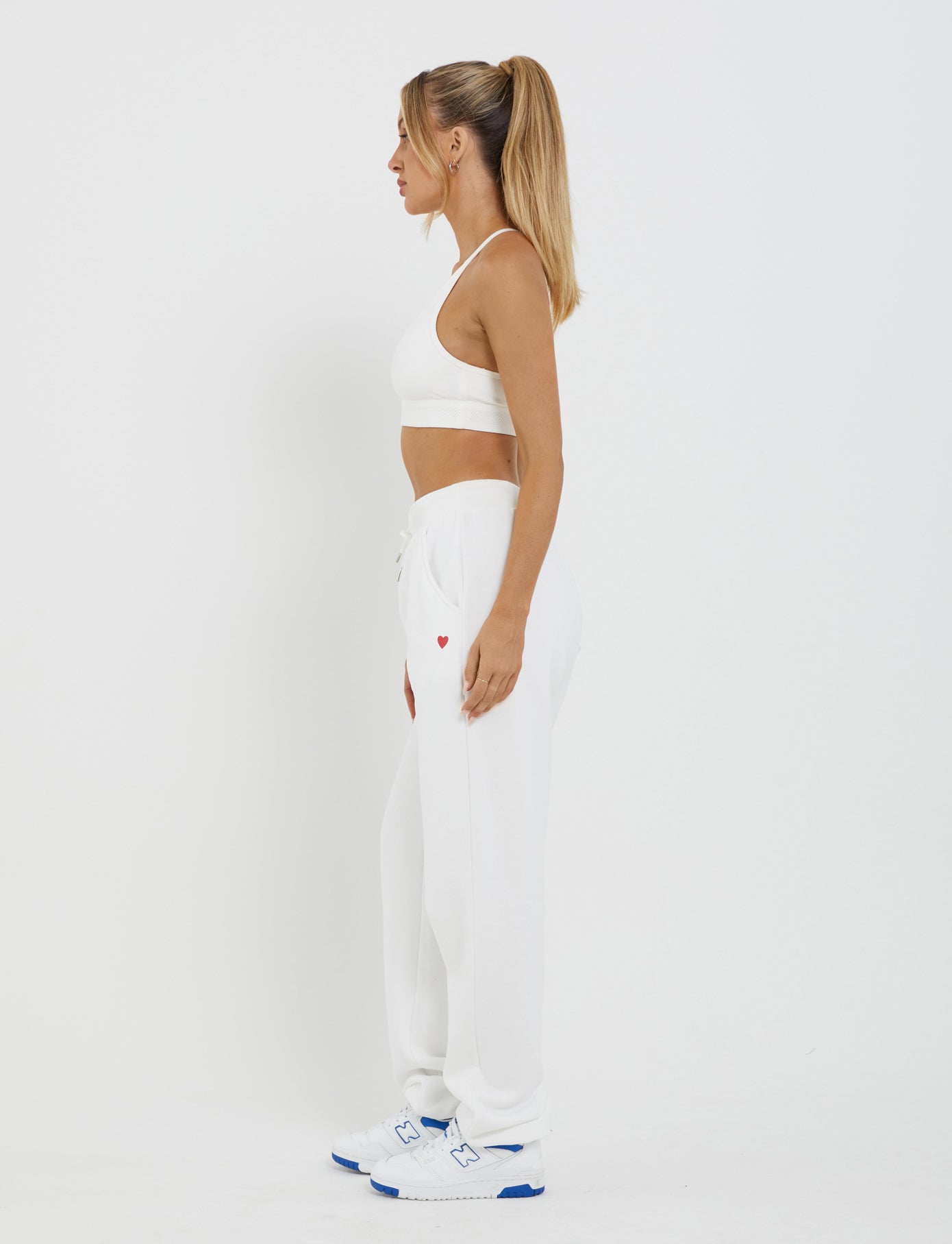 A woman with a high ponytail is standing in profile against a plain background. She is wearing a white sports bra, the LUMEN PANT - WHITE by TIGERMIST featuring an oversized trackpant silhouette and a small red heart near the pocket, and white sneakers with blue accents.