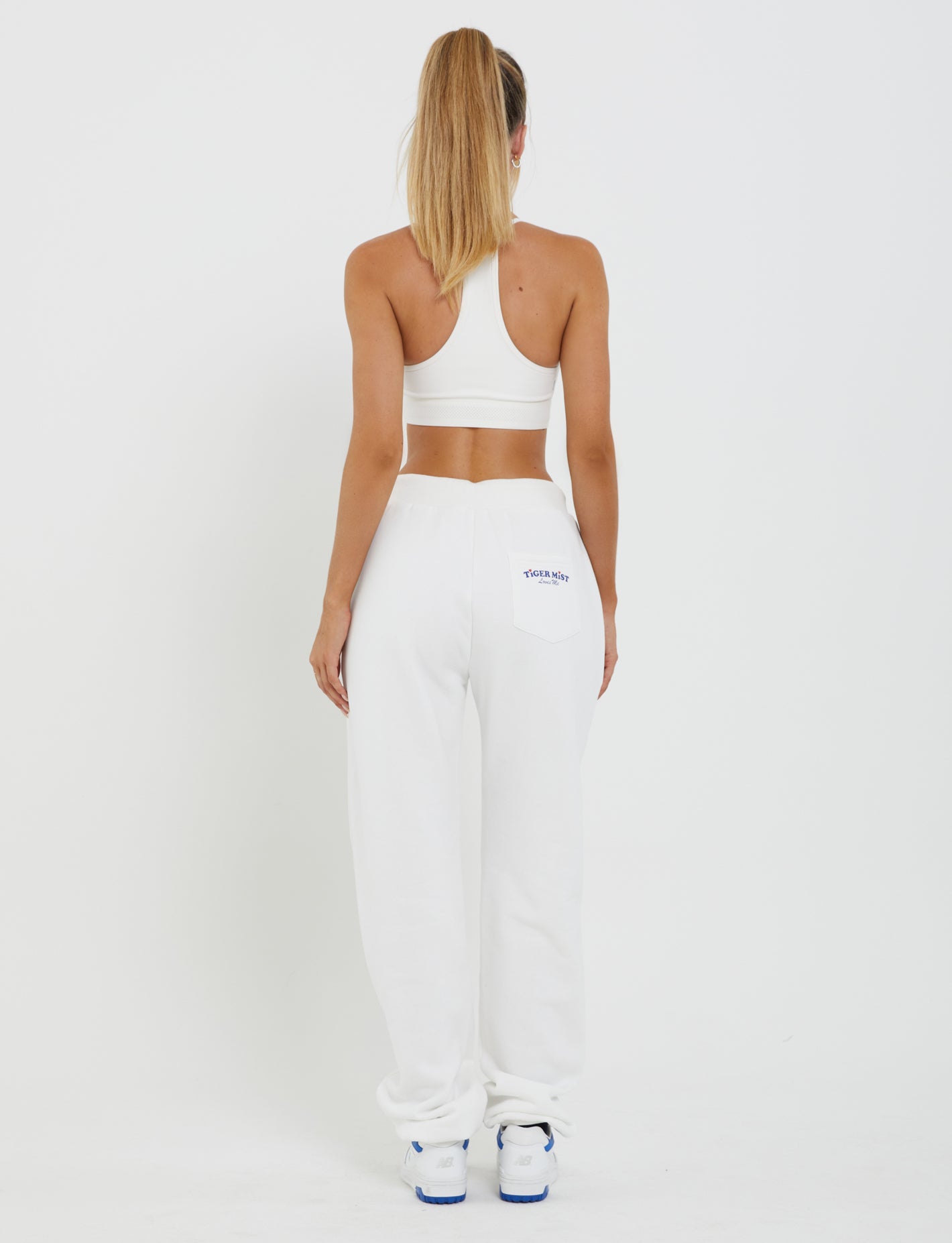 A woman stands with her back to the camera, dressed in the TIGERMIST LUMEN PANT - WHITE and a matching white sports bra. She has long blonde hair styled in a high ponytail and is positioned against a plain white backdrop. Blue text adorns the back pocket of her mid-rise loungewear pants. Her outfit is completed with white sneakers featuring blue accents.
