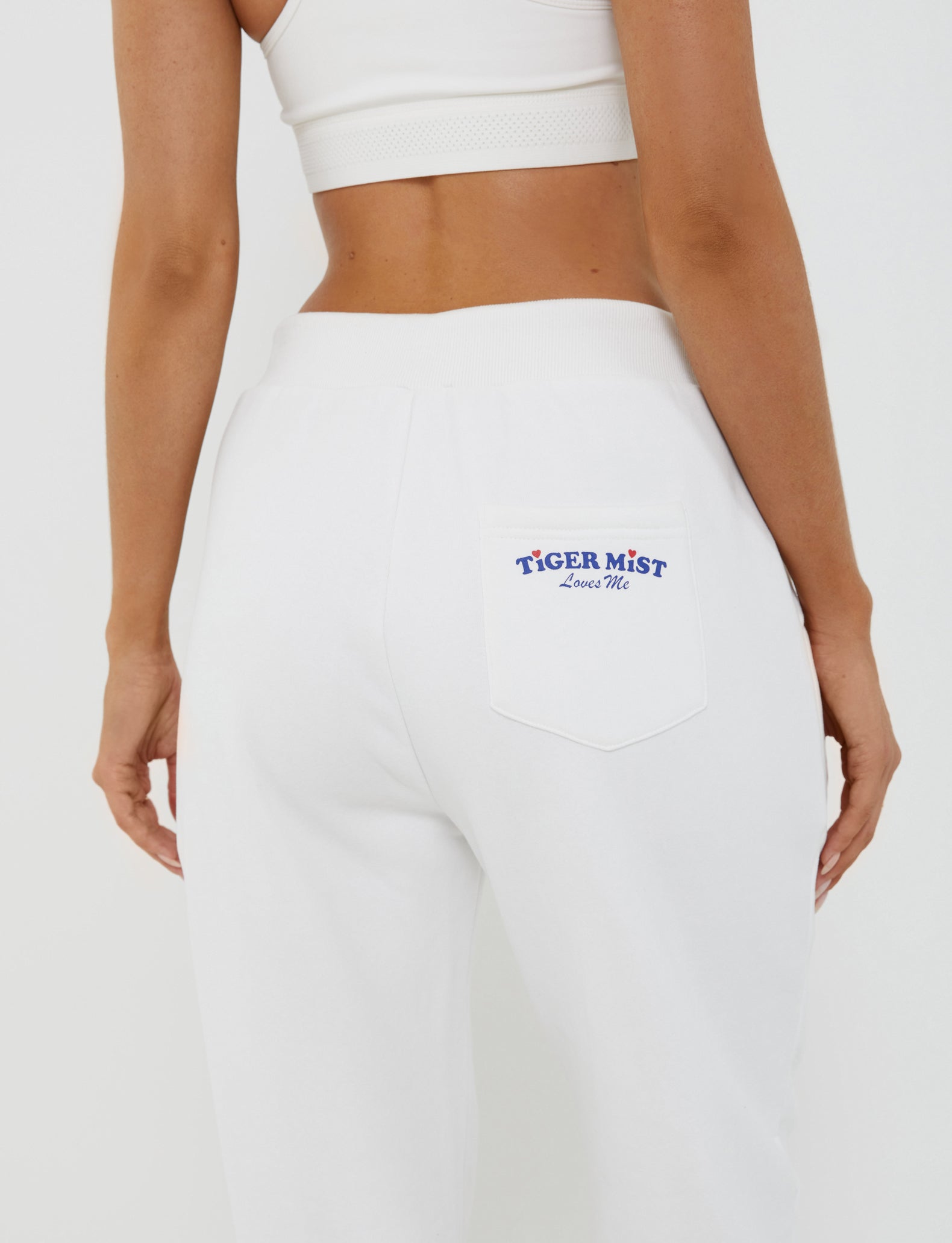 A person is wearing the white LUMEN PANT from TIGERMIST, featuring "Tiger Mist Loves Me" printed in blue on the back pocket. They are also sporting a white crop top. The image centers on their back and lower torso against a plain white background, emphasizing the mid-rise waist of the outfit.