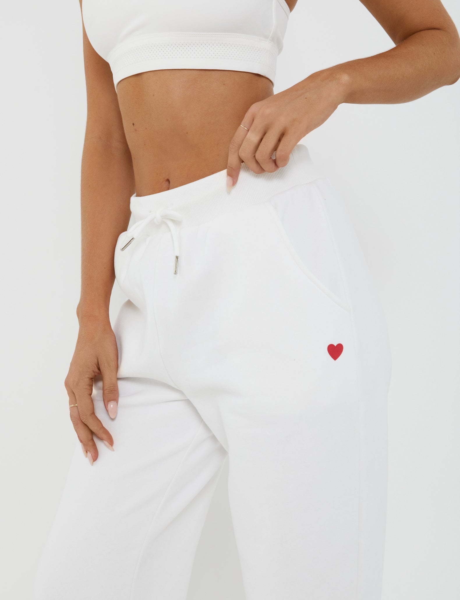 A person wearing a white sports bra and the TIGERMIST LUMEN PANT - WHITE, which features a small red heart on the left pocket, stands against a plain white background. The mid-rise waist pants boast an oversized trackpant silhouette, and the person is pointing to the red heart on the pocket.