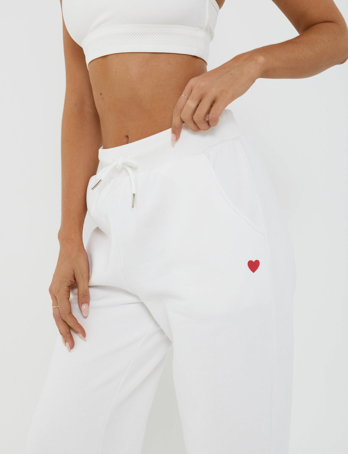 A person wearing a white sports bra and the TIGERMIST LUMEN PANT - WHITE, which features a small red heart on the left pocket, stands against a plain white background. The mid-rise waist pants boast an oversized trackpant silhouette, and the person is pointing to the red heart on the pocket.