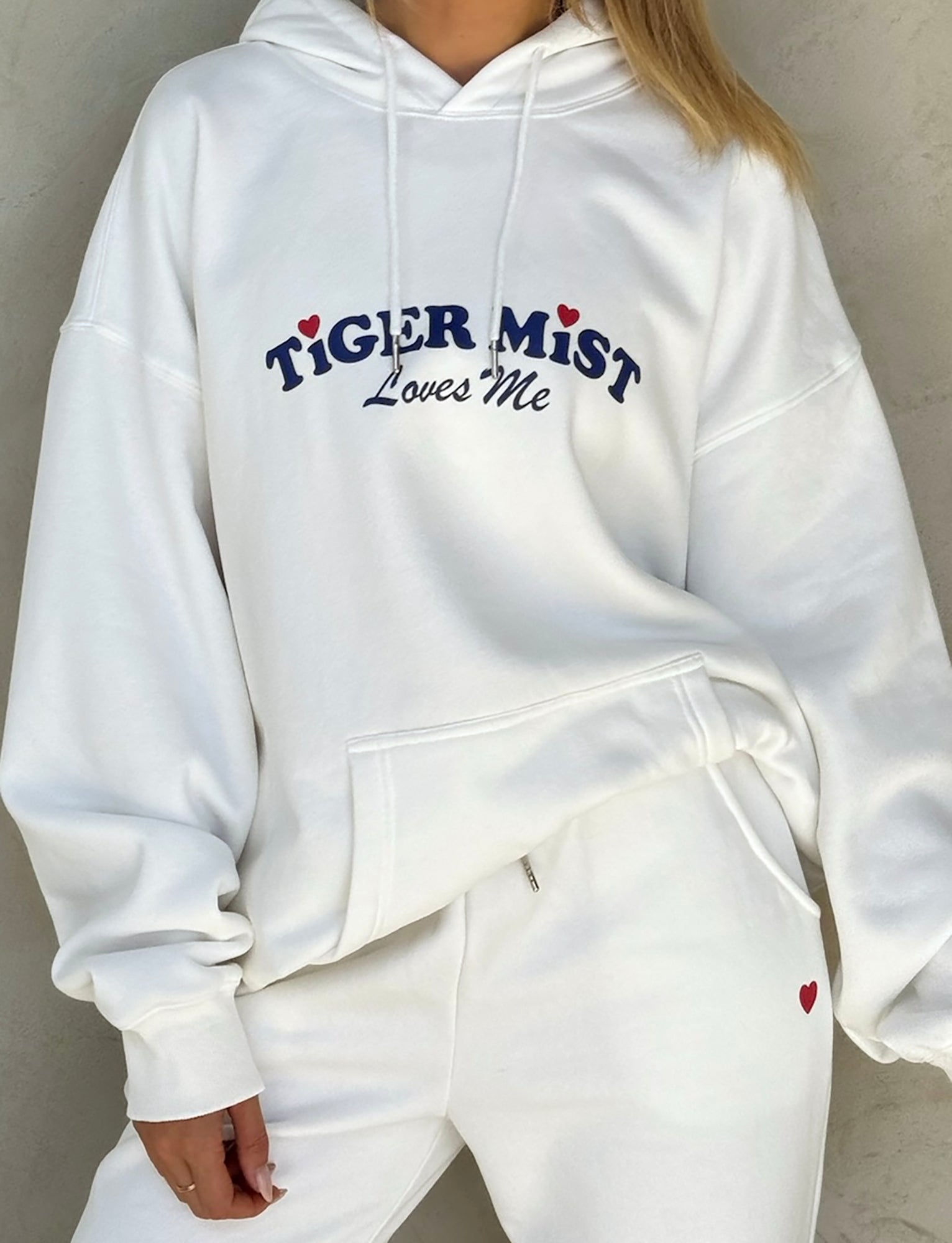 A person is wearing an oversized LUMEN HOODIE - WHITE loungewear from TIGERMIST, featuring a kangaroo pocket and the text "TIGER MIST Loves Me" in blue with small red heart accents on the front. They are also wearing matching sweatpants that have a small red heart near the pocket.