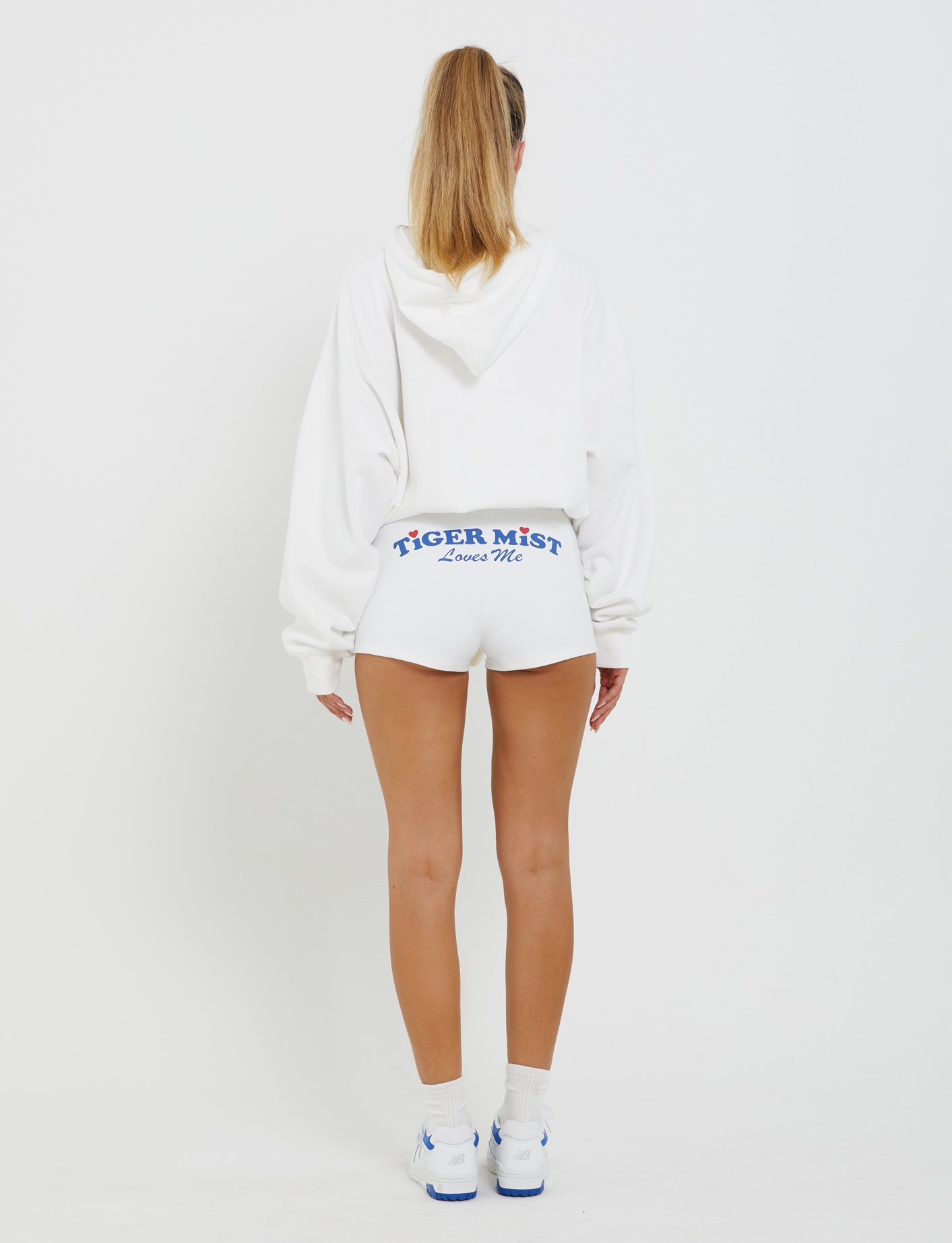 A person with long hair in a high ponytail is standing with their back to the camera. They're wearing an oversized TIGERMIST LUMEN HOODIE in white, white athletic shorts reading "TIGER MIST Loves Me" on the back, white socks, and white sneakers with blue accents.