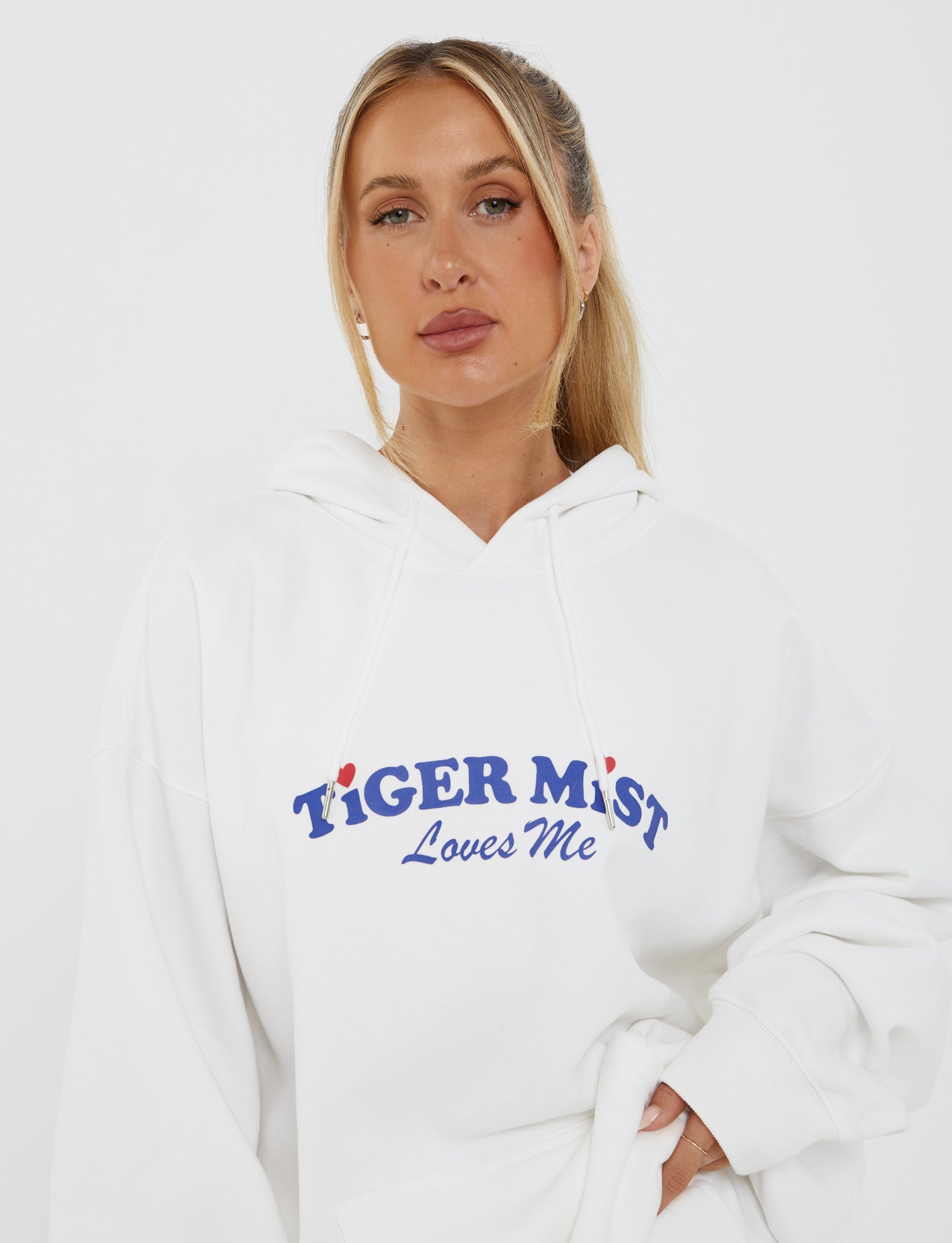 A person with long blonde hair tied back is wearing the LUMEN HOODIE - WHITE from TIGERMIST, which features an oversized silhouette. The hoodie displays the phrase "TIGER MIST Loves Me" printed in blue letters with small red hearts. The person's expression is neutral, and they stand against a plain white background.