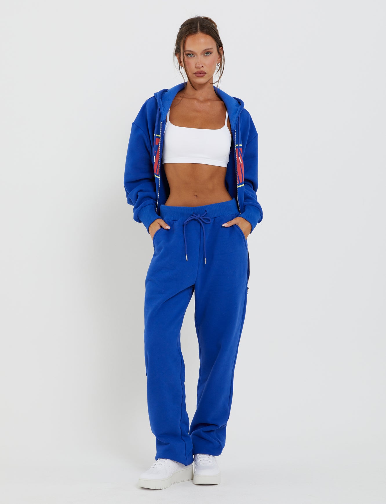 A woman stands against a plain background, wearing the LUMEN PANT - BLUE track pants from TIGERMIST with a matching blue jacket and a white sports bra underneath. Her hands are in the pockets of her track pants, and she is also wearing white sneakers. Her hair is pulled back, and she has a focused expression on her face.