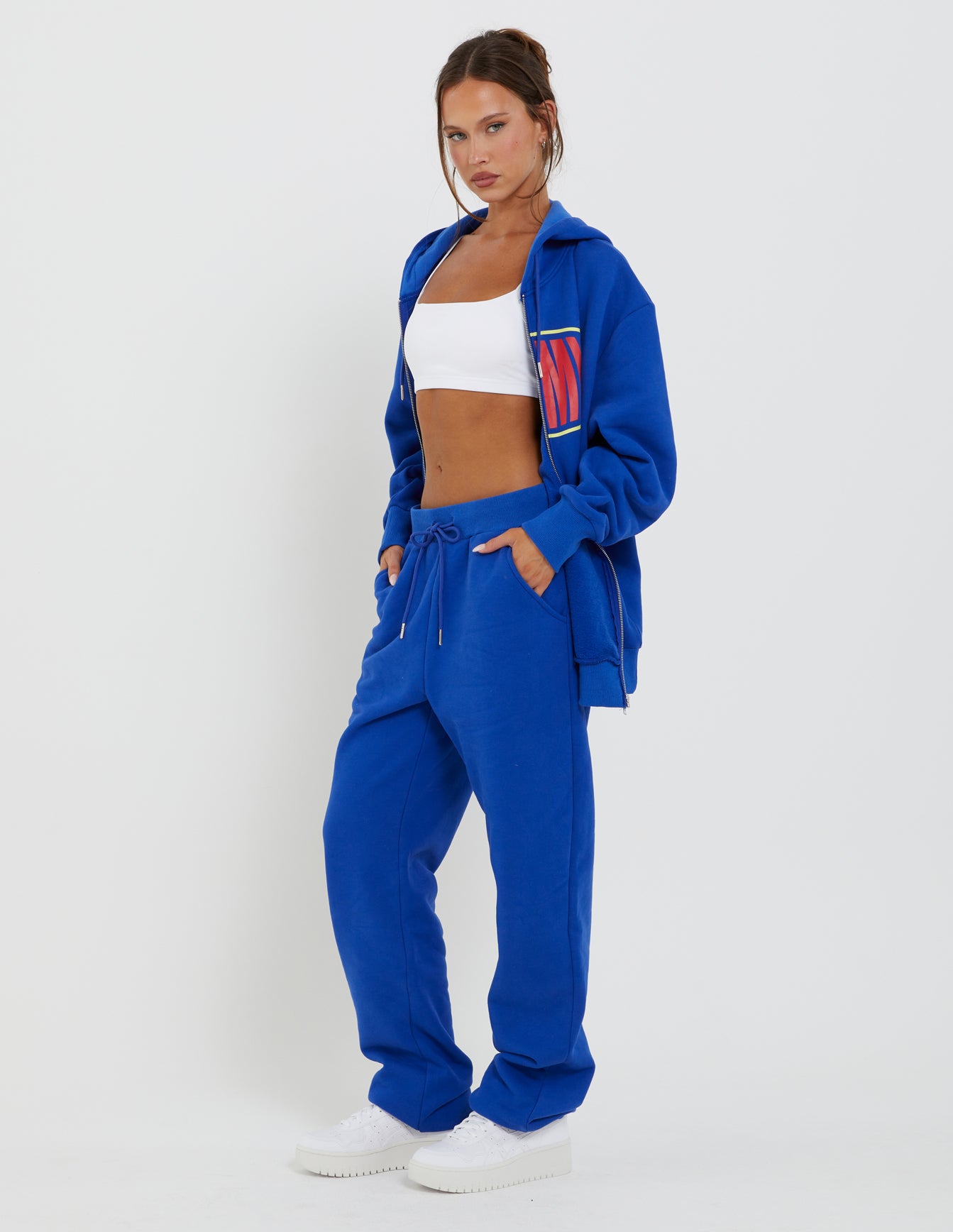 A person with a serious expression wears a blue LUMEN PANT from TIGERMIST, paired with an unzipped blue hoodie, white crop top, and white sneakers. Their hands are in the pockets of the LUMEN PANT as they stand against a plain white background.