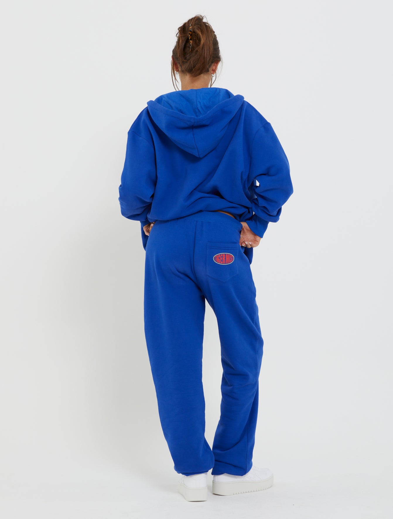 A person stands with their back to the camera, wearing a bright blue hoodie and matching LUMEN PANT - BLUE from TIGERMIST. Their hair is tied up in a casual bun. The pants feature a small, circular red and blue logo patch on the right back pocket. The background is plain white.