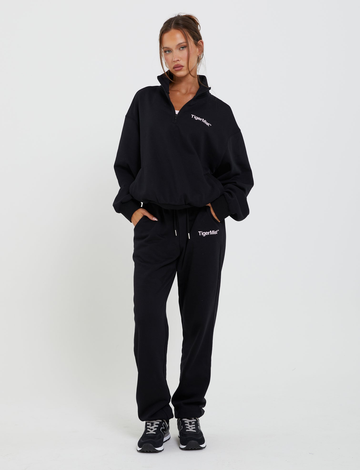 A woman stands against a plain white background wearing the TIGERMIST DELFINA PANT - BLACK tracksuit, which has "TIGERMIST" printed on the top left of both the jacket and pants. She has her hands in her pockets and is also wearing black athletic shoes. Her hair is tied back neatly.