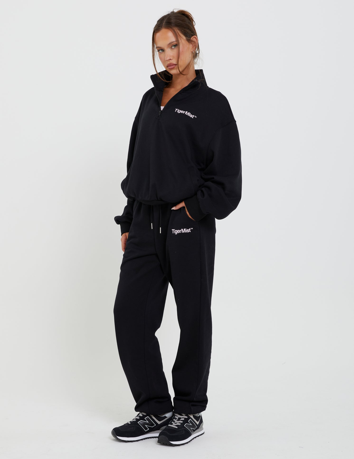 A person stands against a white background wearing a black quarter-zip sweatshirt and matching DELFINA PANT - BLACK from TIGERMIST. "TIGERMIST" is printed on both pieces. With hands in the joggers' pockets, black sneakers, and hair tied back, they present a sleek and stylish look.
